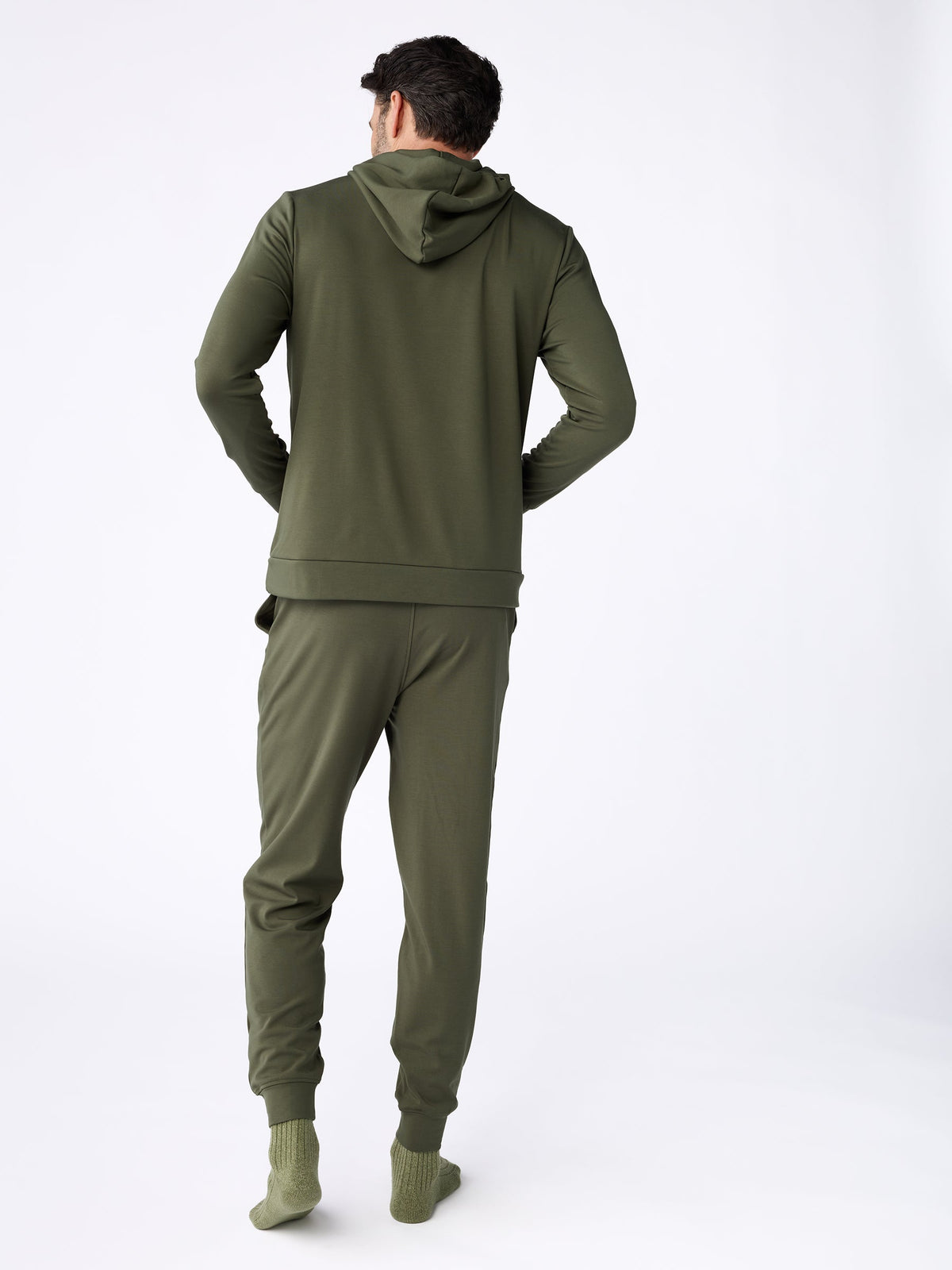 A person is modeling the Cozy Earth Men's Ultra-Soft Bamboo Pullover Crew and matching sweatpants in olive green, with their back to the camera. They are also wearing green socks in a plain white studio setting. |Color:Olive