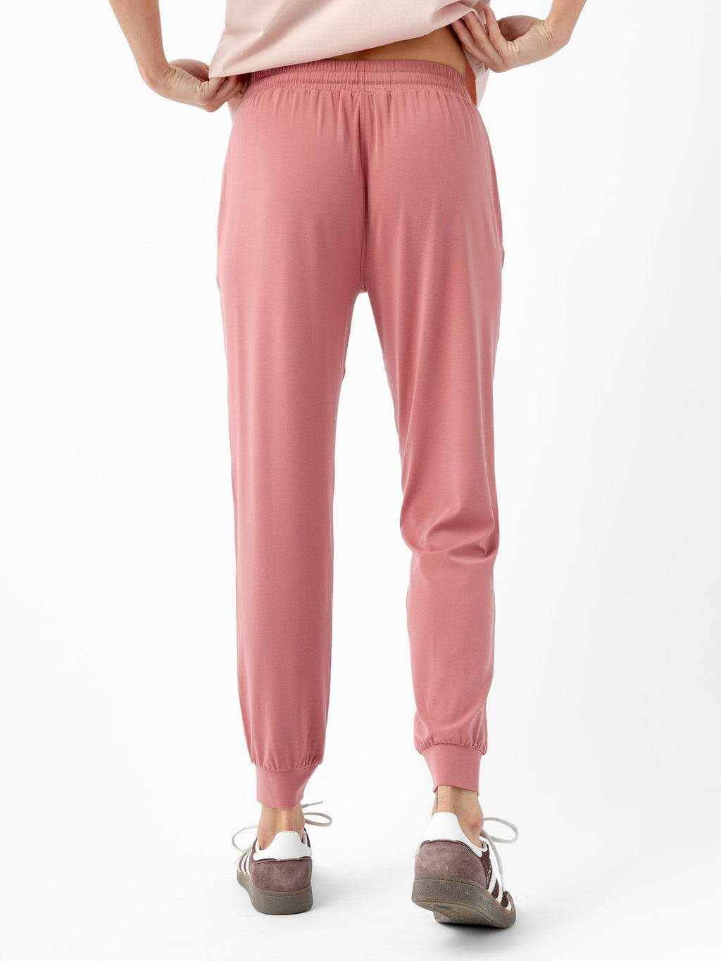 A person standing with their back to the camera, wearing Cozy Earth's rose-colored Women's Studio Jogger and an off-white top. They are also wearing brown and white sneakers. The person's hands are positioned on their hips against a plain white background. |Color:Rouge