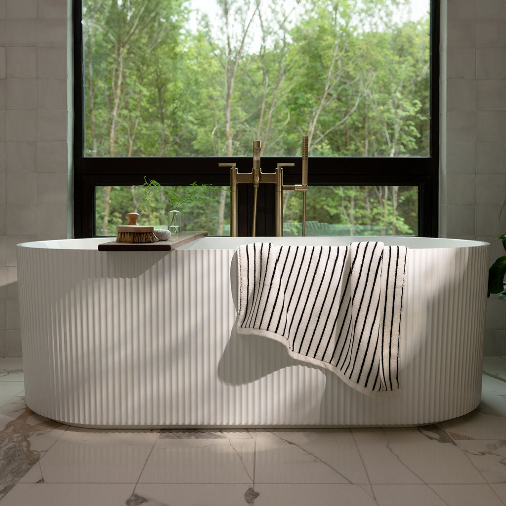 A serene bathroom features a white freestanding bathtub with a Luxe Bath Towel from Cozy Earth draped over the side. Behind the tub, a large window showcases a lush green forest, allowing natural light to flood the space. The floor is made of white marble with subtle grey veins. |Color:Black Stripe