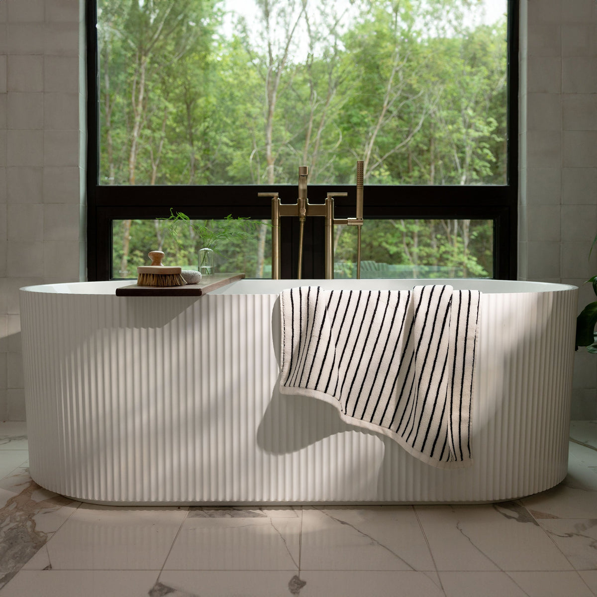 A serene bathroom features a white freestanding bathtub with a Luxe Bath Towel from Cozy Earth draped over the side. Behind the tub, a large window showcases a lush green forest, allowing natural light to flood the space. The floor is made of white marble with subtle grey veins. |Color:Black Stripe