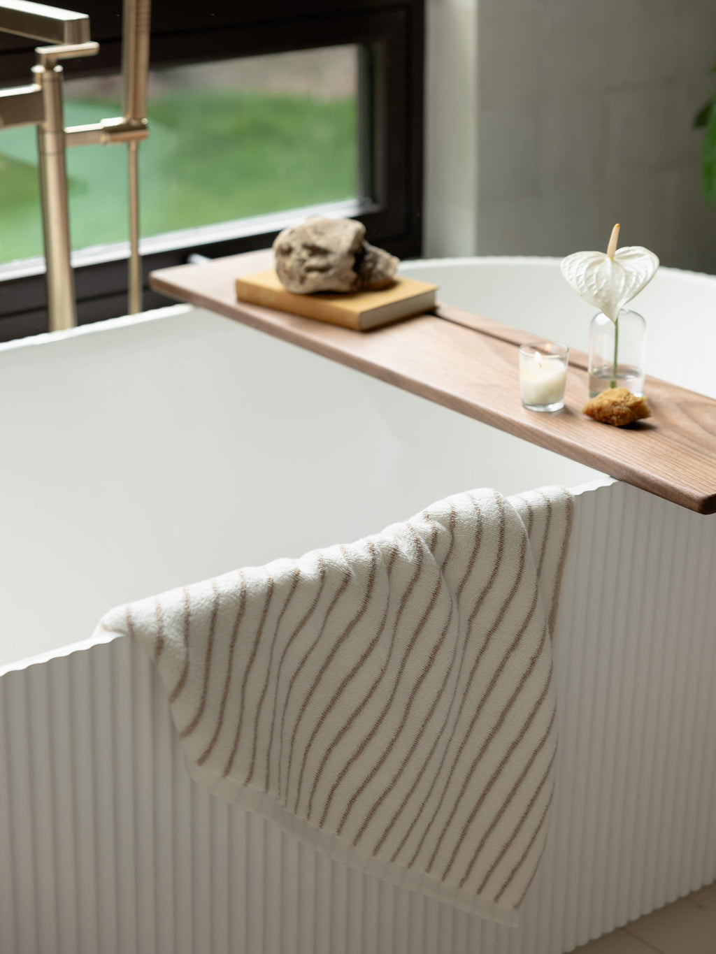 A modern white bathtub with a textured exterior stands near a window. A wooden tray spans the tub, holding two glass vases with plants, a candle, and a decorative stone. Draped over the tub's edge is a Luxe Bath Towel featuring thin, wavy brown stripes from Cozy Earth. |Color:Sand Stripe