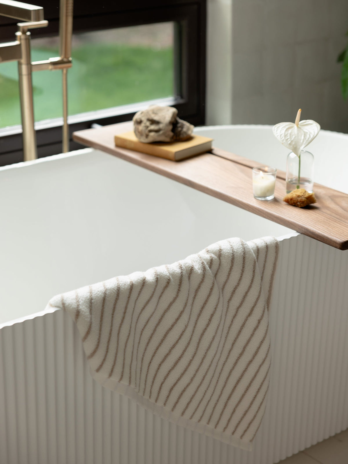 A modern white bathtub with a textured exterior stands near a window. A wooden tray spans the tub, holding two glass vases with plants, a candle, and a decorative stone. Draped over the tub's edge is a Luxe Bath Towel featuring thin, wavy brown stripes from Cozy Earth. |Color:Sand Stripe