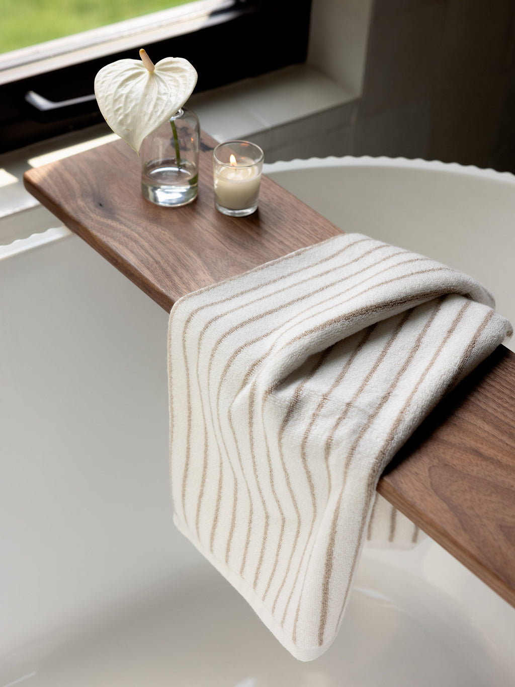 A cozy bathroom setting featuring a wooden bathtub tray with a white anthurium flower in a clear vase, a lit candle, and a folded Luxe Hand Towel by Cozy Earth, striped in white and beige, draped over the tray. The bathtub is white and positioned near a window with greenery outside. |Color:Sand Stripe
