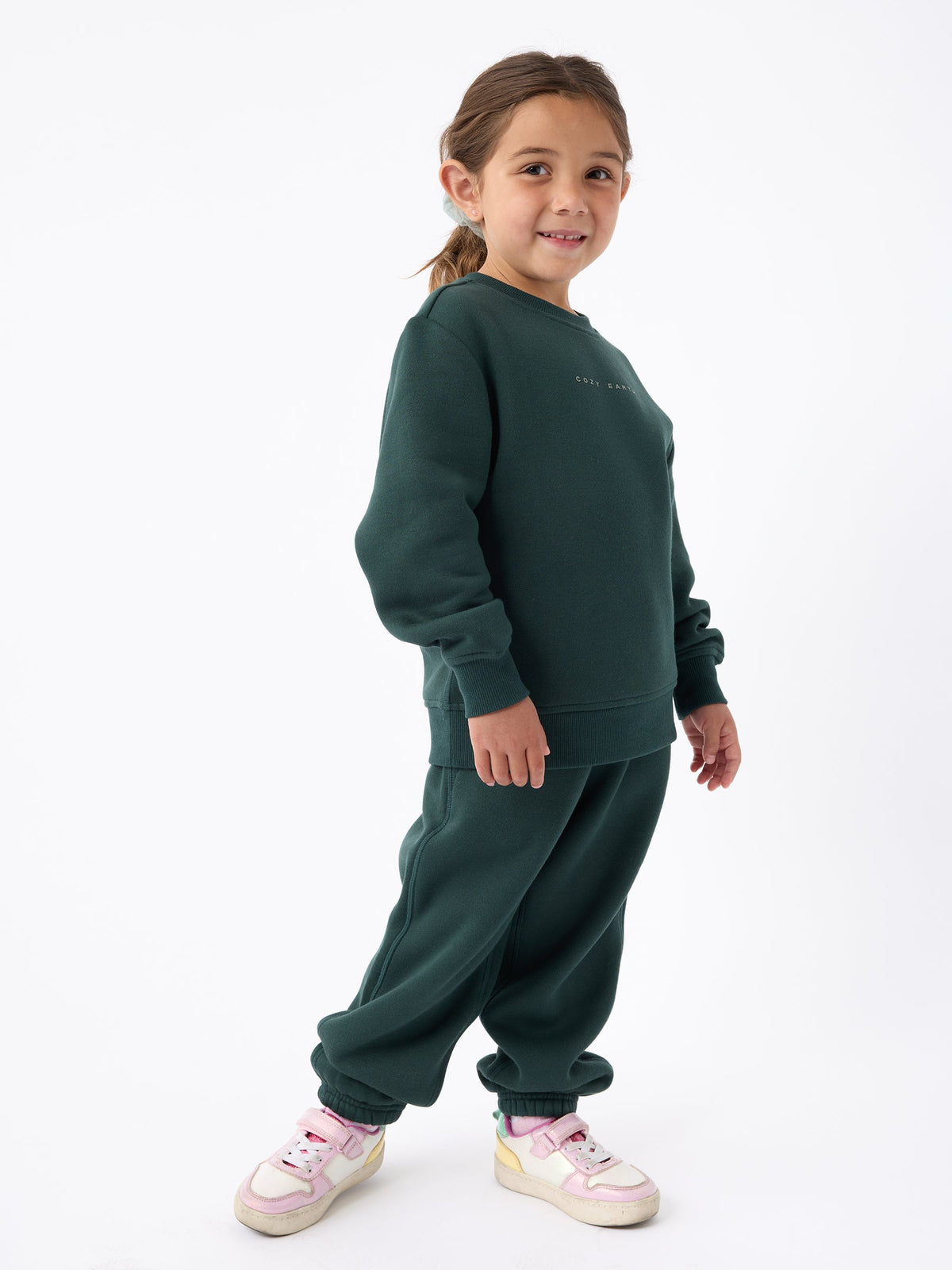 A young child with brown hair stands against a white background, smiling. They are wearing a dark green Kid's CityScape Crewneck by Cozy Earth, paired with sweatpants and pink and white sneakers. |Color:Spruce