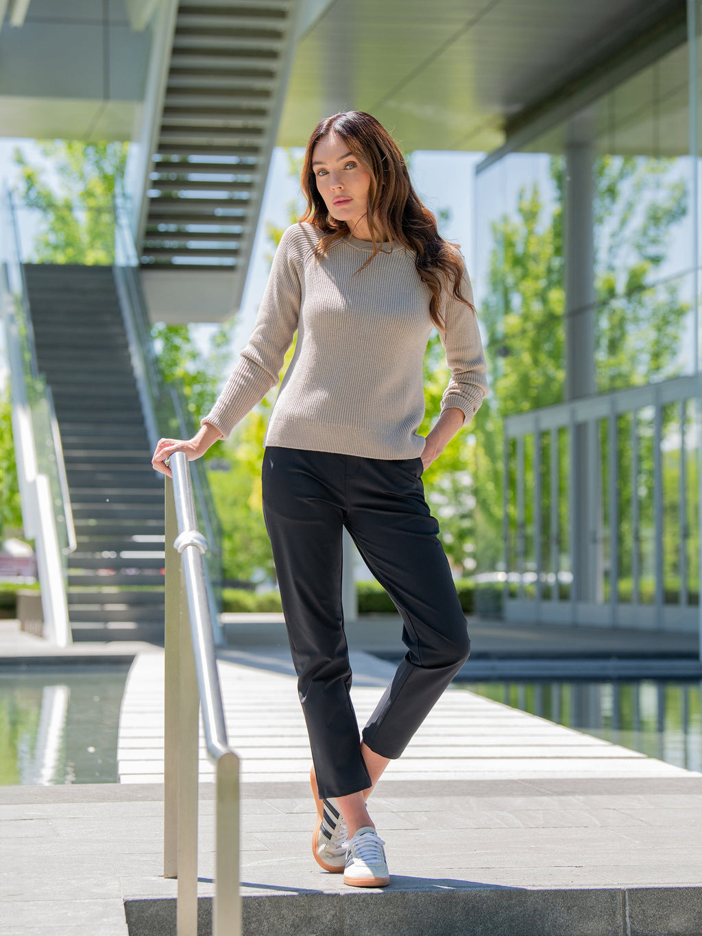 A person in Cozy Earth's Women's Classic Crewneck and black pants stands outdoors on a walkway by a railing. They have one hand on the railing and the other in their pocket. They are under a modern building structure featuring glass and metal elements, with greenery visible around. |Color:Taupe