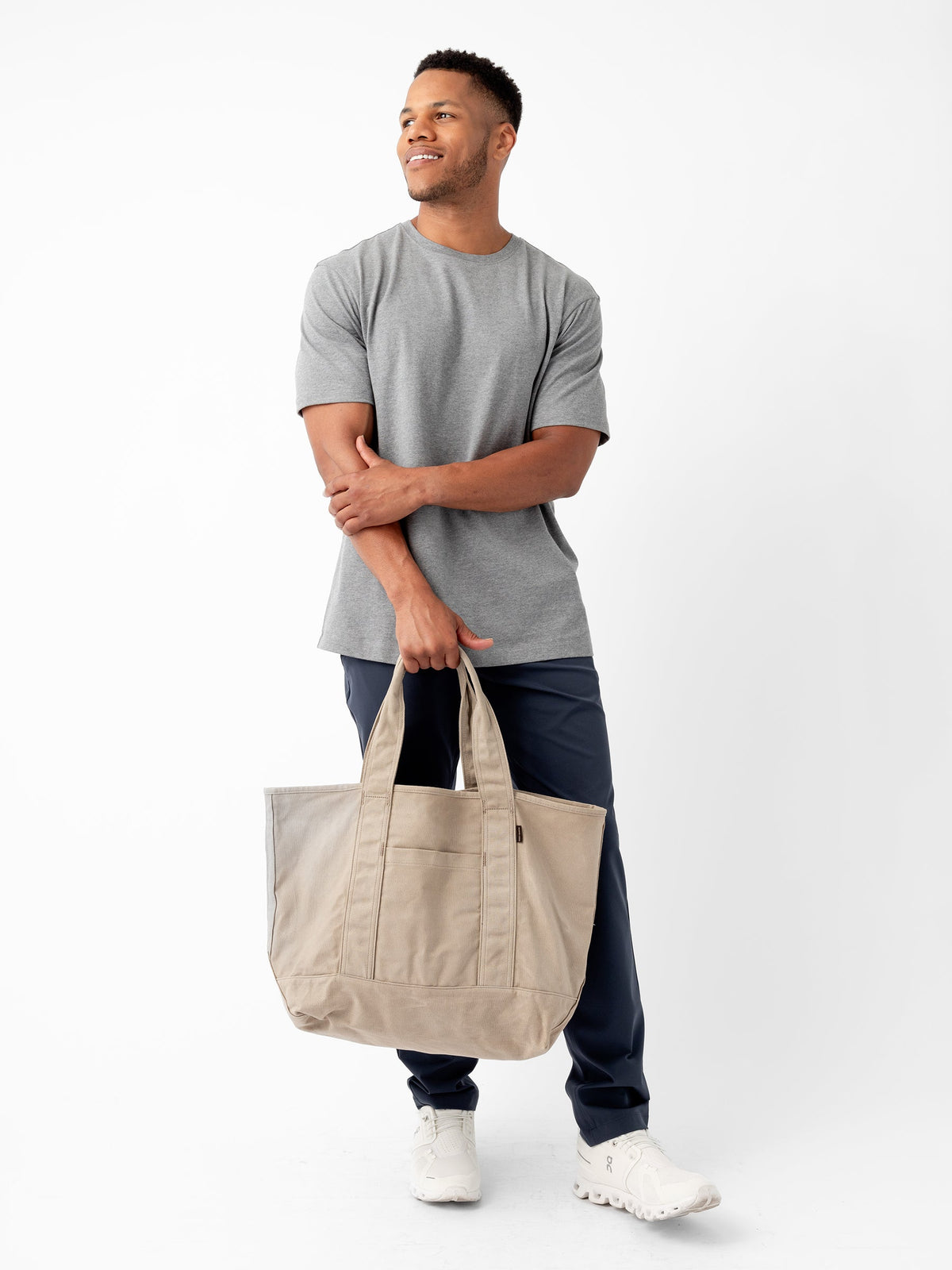 A man stands against a plain white background, smiling slightly while looking off to the side. He is wearing a gray T-shirt, dark blue pants, and white sneakers, and is holding the Waxed Canvas Tote from Cozy Earth. His arms are crossed casually as he carries the bag. |Color:Taupe