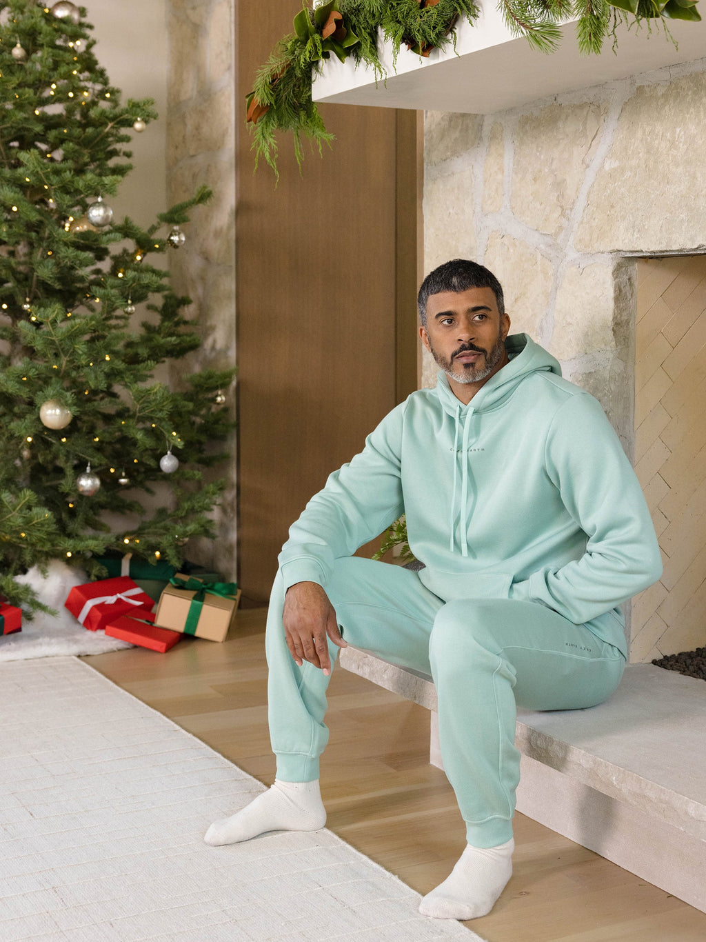 A man in a mint-green CityScape Jogger by Cozy Earth sits by a stone fireplace adorned with greenery. A decorated Christmas tree and wrapped gifts are nearby, creating a festive atmosphere. The room has a cozy and relaxed holiday vibe. |Color:Wintergreen