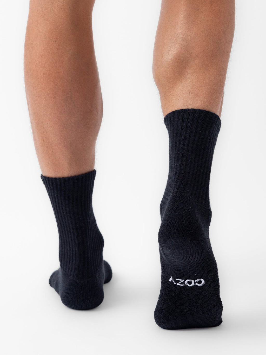 A person stands on tiptoe, viewed from behind, wearing black Essential Calf Socks by Cozy Earth. "COZY" is printed in white on the sole of the right sock. The background is plain white. |Color:Black