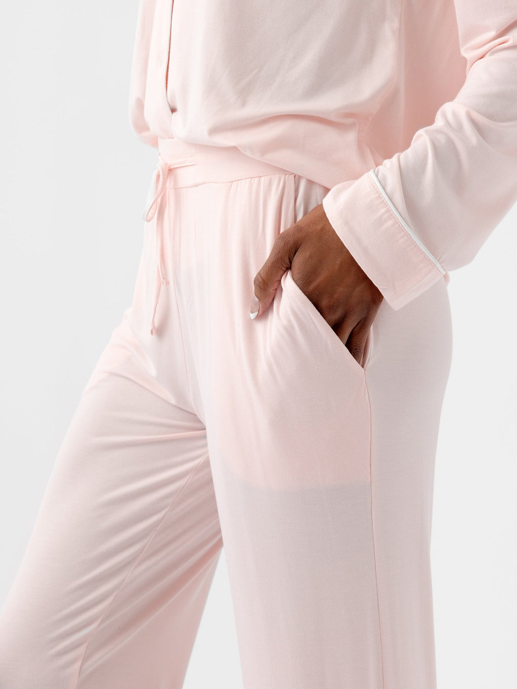 A person dressed in HIDE's Women's Stretch-Knit Bamboo Pajama Pant TALL in light pink is shown from the shoulders down, with their left hand in the pocket of their pants. The soft and loose-fitting outfit features a long-sleeve top and drawstring pants against a plain white background. |Color:Blossom