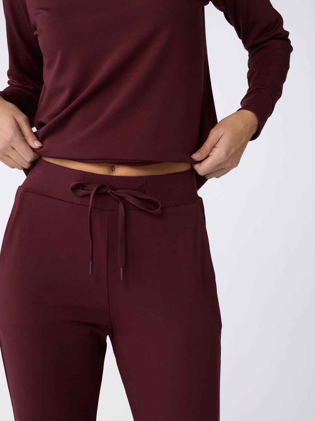 A person dressed in a maroon long-sleeve top and Cozy Earth's Women's Bamboo Jogger Pant with a matching drawstring is adjusting their shirt against a plain white background. |Color:Burgundy