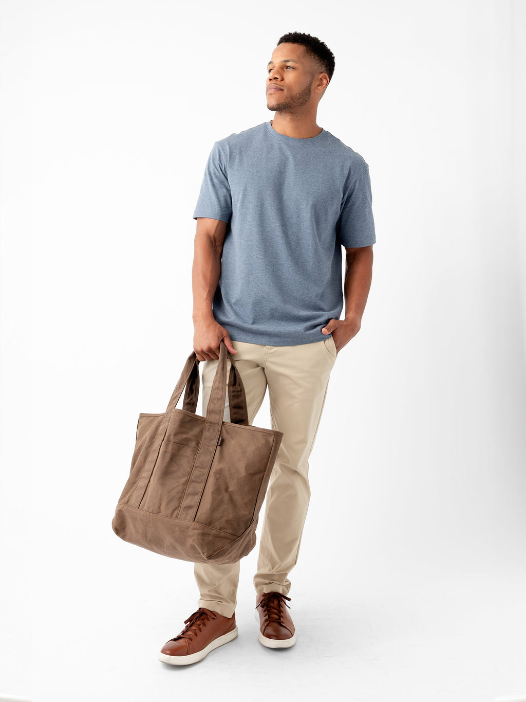 A man stands against a plain white background. He is wearing a blue T-shirt, beige pants, and brown sneakers. He holds a large Waxed Canvas Tote from Cozy Earth in his left hand and has his right hand in his pants pocket. He looks slightly to the left with a neutral expression. |Color:Cedar