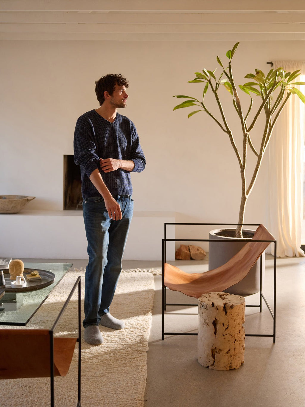 A person stands in a minimalist living room featuring a large plant, a unique wooden chair, and contemporary furniture. Dressed in jeans and a dark shirt by Cozy Earth, they gaze to the side in the well-lit space of neutral tones. |Color:Charcoal