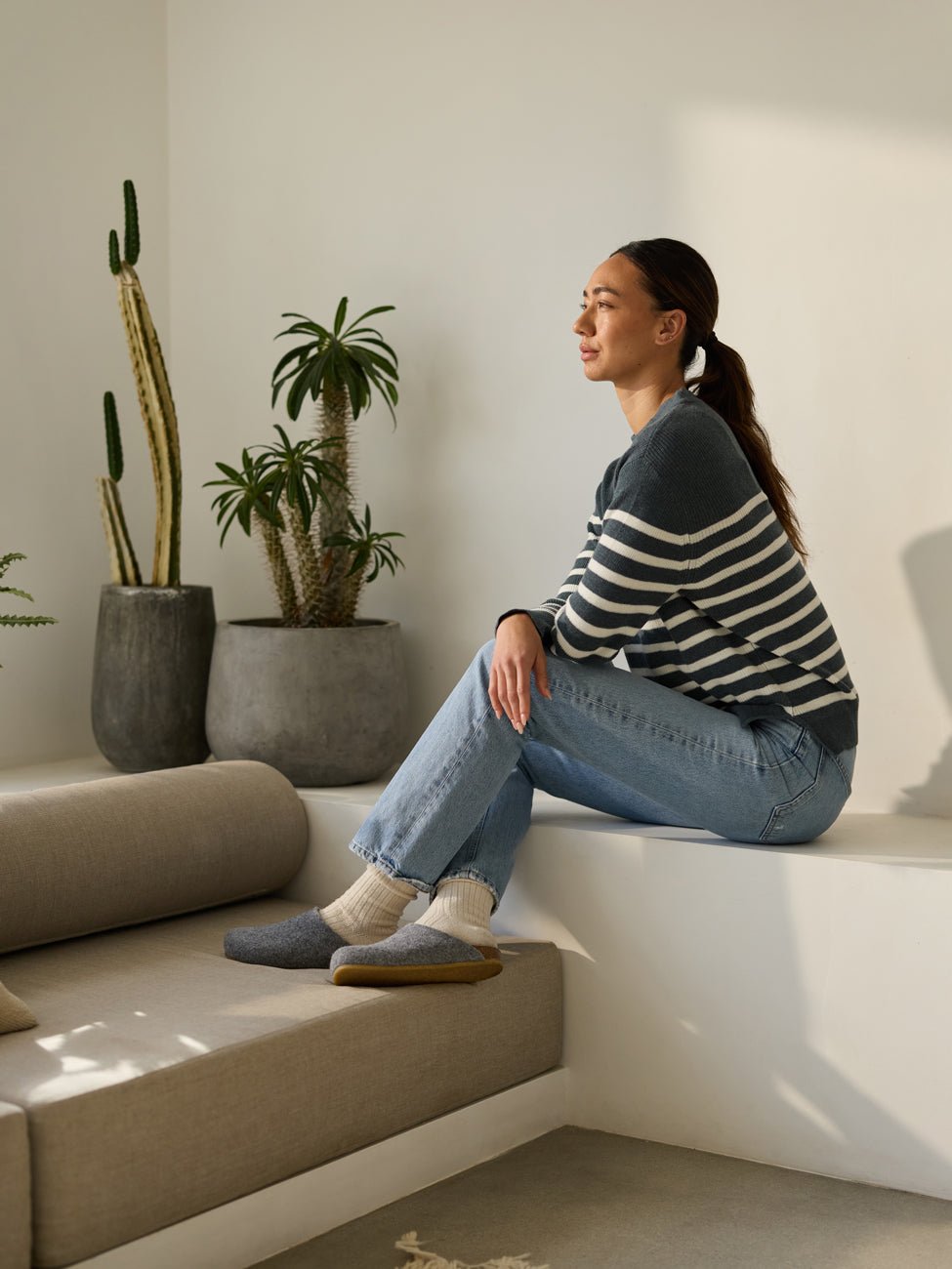 A person wearing a striped sweater and jeans sits on a ledge in a sunlit room, looking to the side with a relaxed expression. They are wearing Lakehouse Clogs by Cozy Earth. Potted plants, including a cactus, decorate the floor in the background. |Color:Charcoal