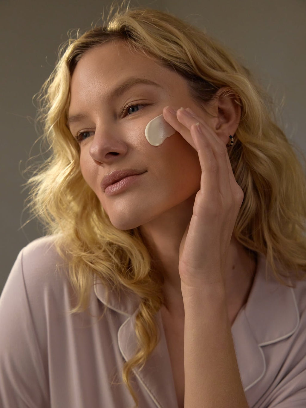 With long, wavy blonde hair, a person applies Cozy Earth's Clay Mask to their cheek, appearing at ease. They're dressed in a pale pink top with a gentle smile. The neutral background emphasizes their skincare ritual. |Size:Full Size
