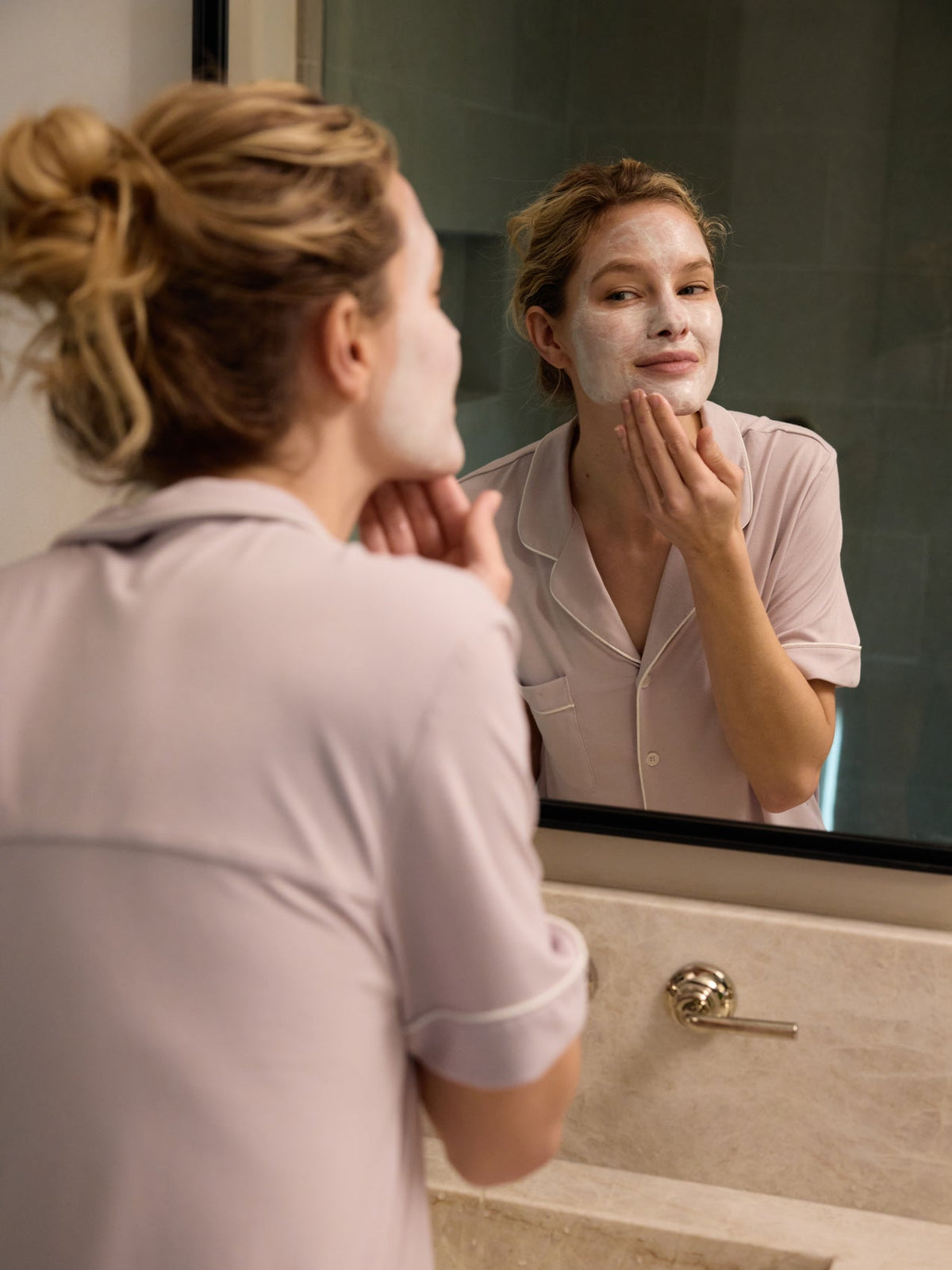 In Cozy Earth pajamas, a person applies a Clay Mask while looking in the bathroom mirror. Their hair is tied back as they stand at the sink, smoothing the mask on their cheek with one hand. |Size:Mini