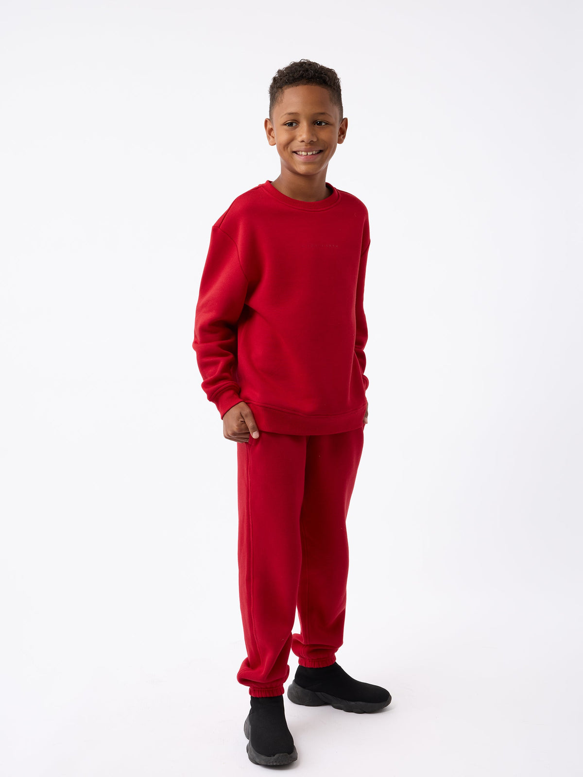 A boy wearing the Kid's CityScape Crewneck by Cozy Earth and black shoes stands smiling against a plain white background. |Color:Crimson