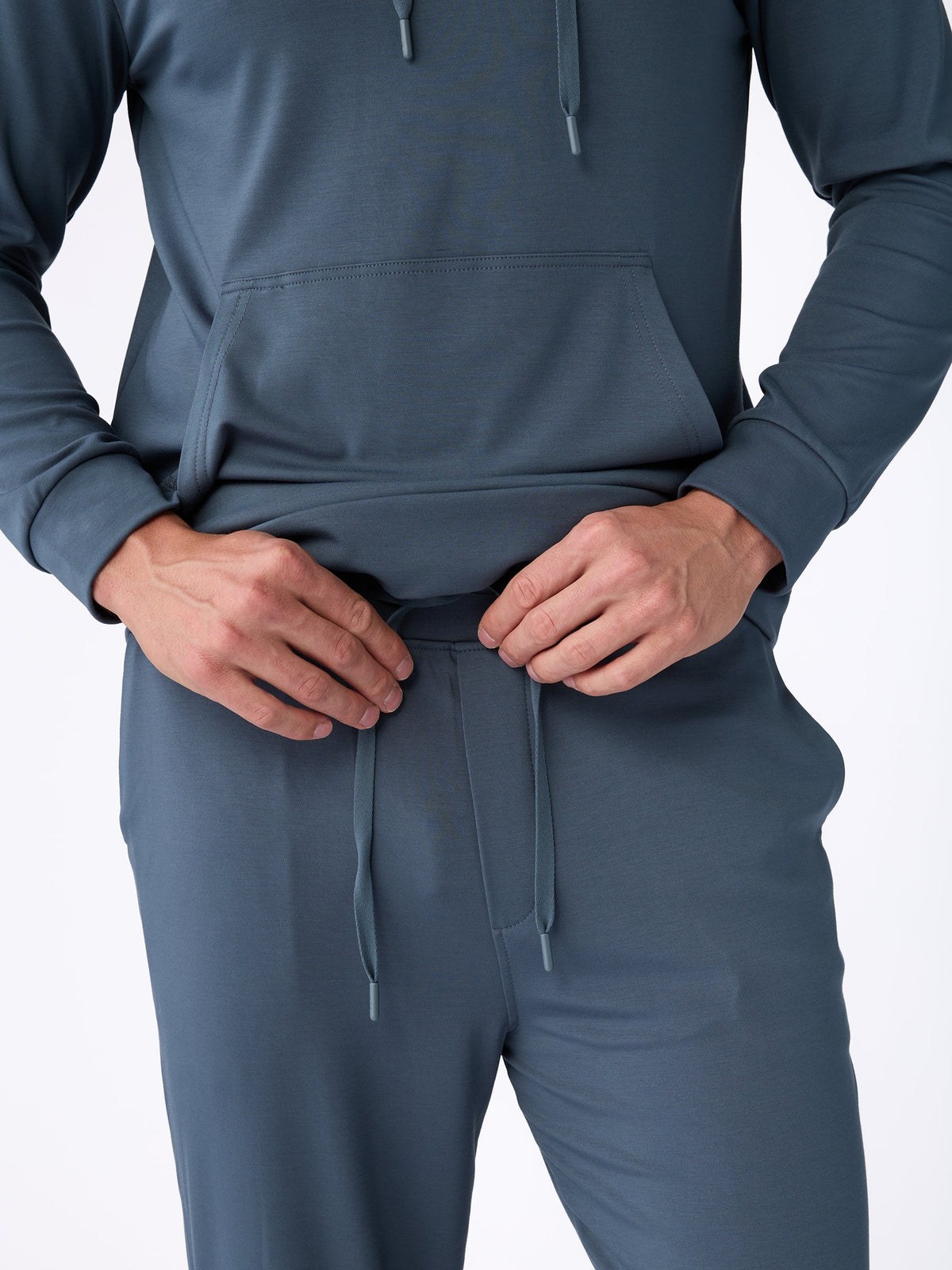 A person wearing a blue tracksuit from Cozy Earth's Men's Ultra-Soft Bamboo Pullover Crew collection adjusts the waistband's drawstring. The outfit includes a hooded sweatshirt with a kangaroo pocket and matching pants, shown against a plain white background. |Color:Dawn
