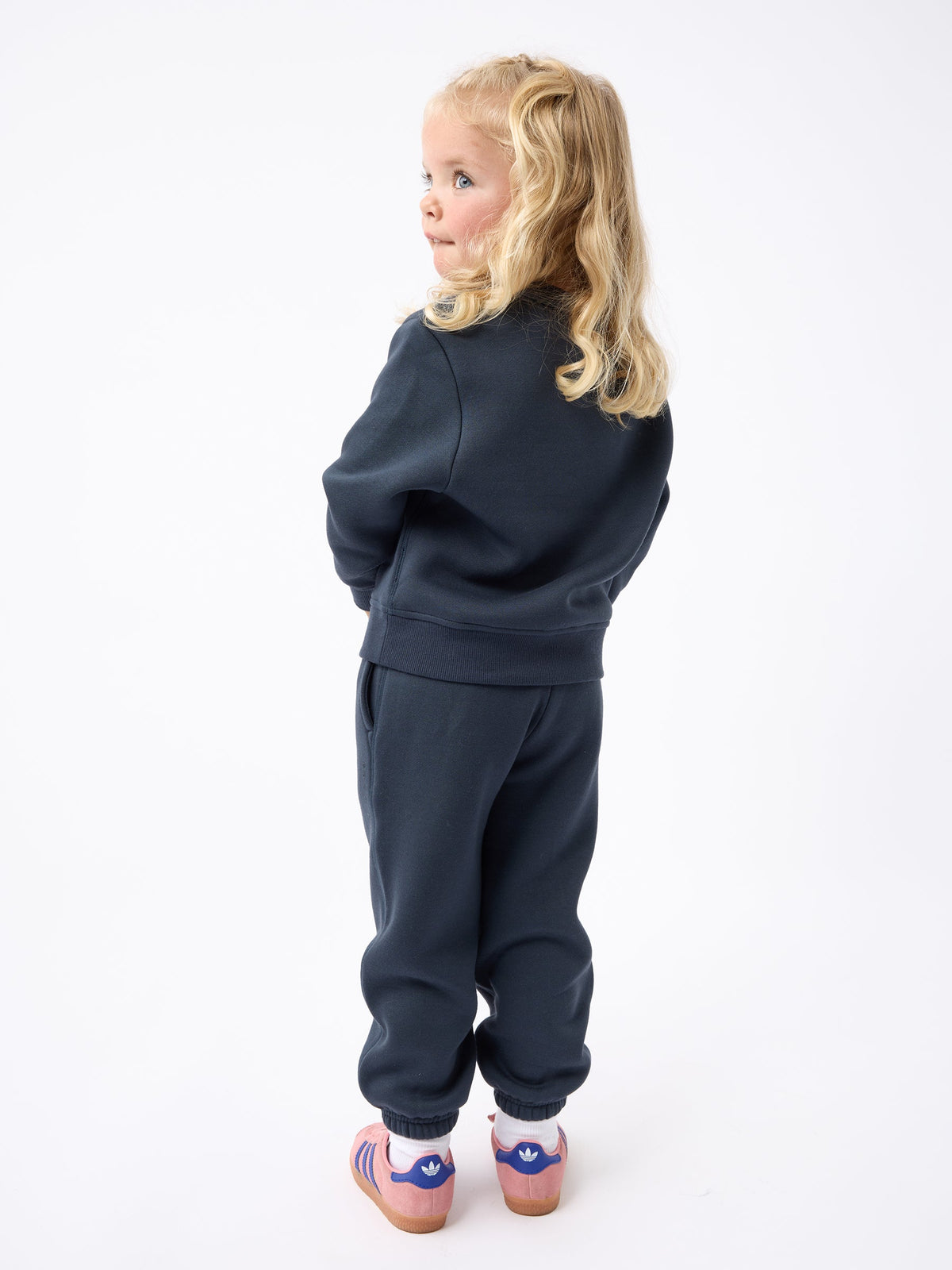 A young child with long blonde hair, viewed from the back and slightly turned to the left, is wearing a Cozy Earth Kid's CityScape Crewneck in dark blue paired with pink striped sandals. The child stands against a plain white backdrop. |Color:Eclipse
