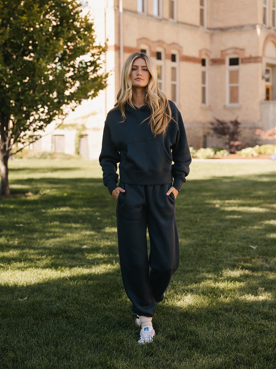 A person with long blonde hair stands on a grassy area in front of a building with brick and stone walls. They are wearing the Women's CityScape Cropped Hoodie by Cozy Earth, paired with dark-colored sweatpants and white sneakers. With their hands in their pockets, they look directly at the camera. A tree is partially visible on the left. |Color:Eclipse