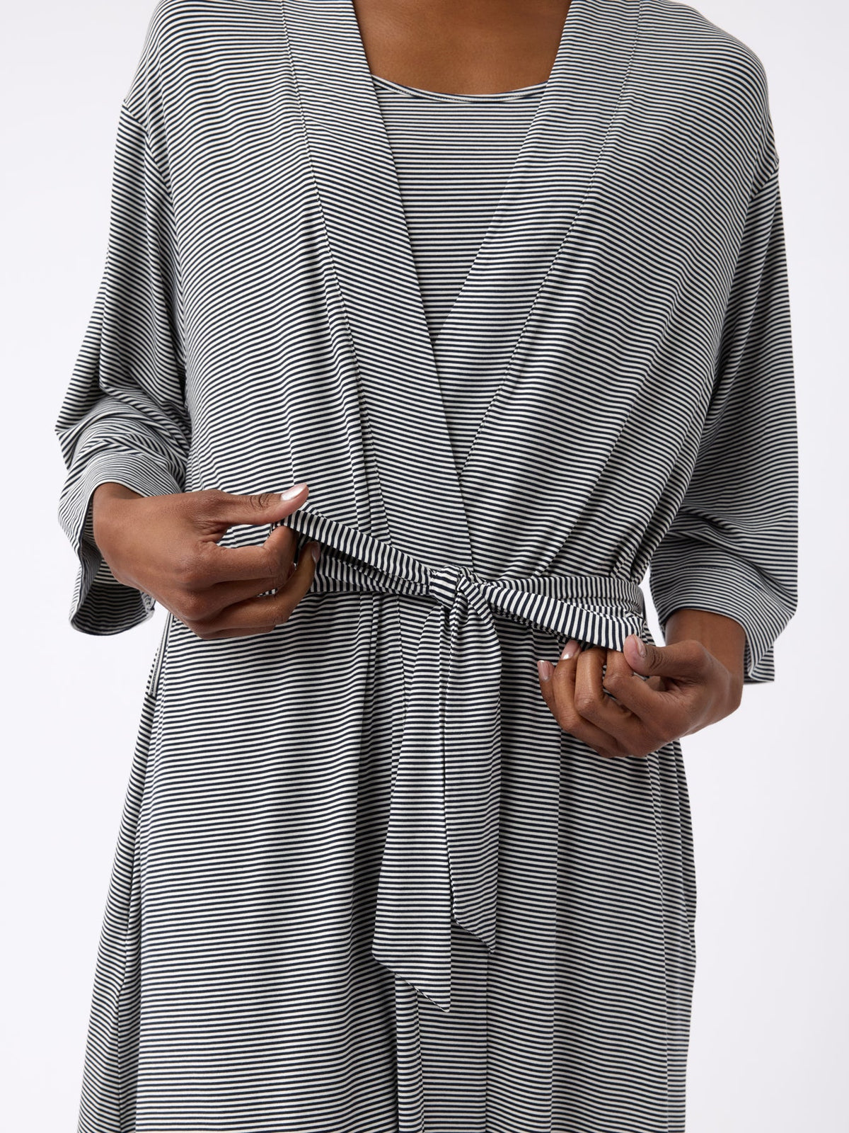 A person adjusts the waist tie of a black and white striped Cozy Earth Women's Bamboo Stretch-Knit Kimono Robe, featuring three-quarter sleeves and a wrap design, with an image centered on their torso and hands. |Color:Eclipse Mini Stripe