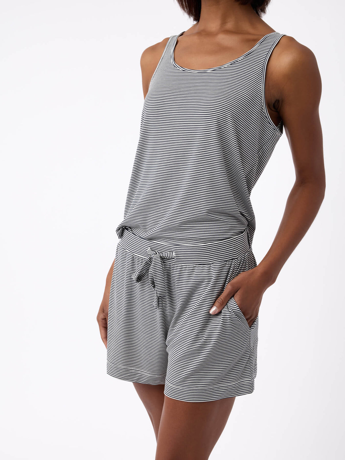 A person in Cozy Earth's Women's Bamboo Stretch-Knit Lounge Tank, a sleeveless gray and white striped romper with pockets, stands against a plain white background. The soft, comfortable fabric complements the casual pose with one hand in a pocket. |Color:Eclipse Mini Stripe