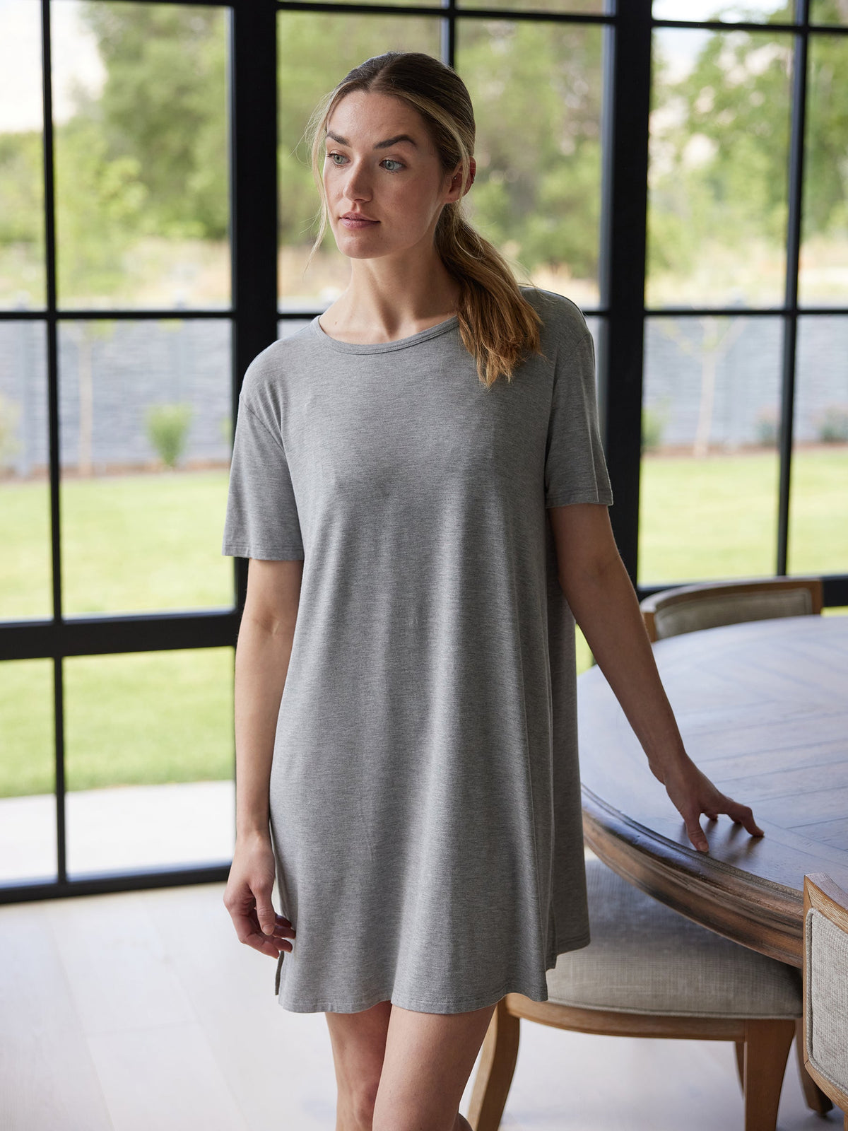 A woman in a Cozy Earth Women's Bamboo Stretch Knit Sleep Dress stands indoors by a wooden table. The gray, short-sleeve dress accentuates the natural light streaming through the large floor-to-ceiling windows, revealing a green lawn in the background. |Color:Heather Grey