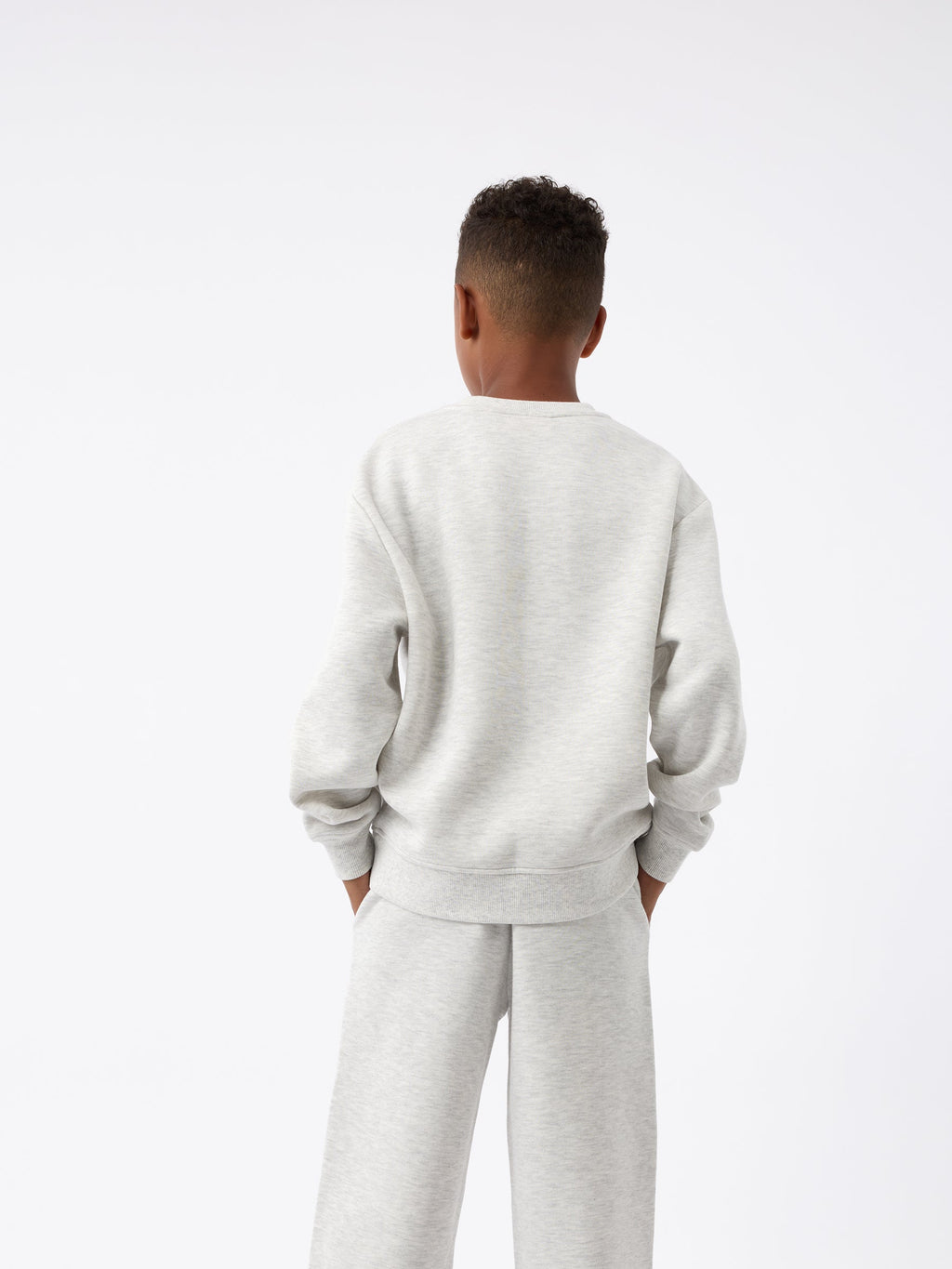 A child dressed in a light gray Kid's CityScape Crewneck from Cozy Earth, accompanied by matching pants, stands with their back to the camera. Their hands are tucked into their pockets against a plain white background. |Color:Heather Grey