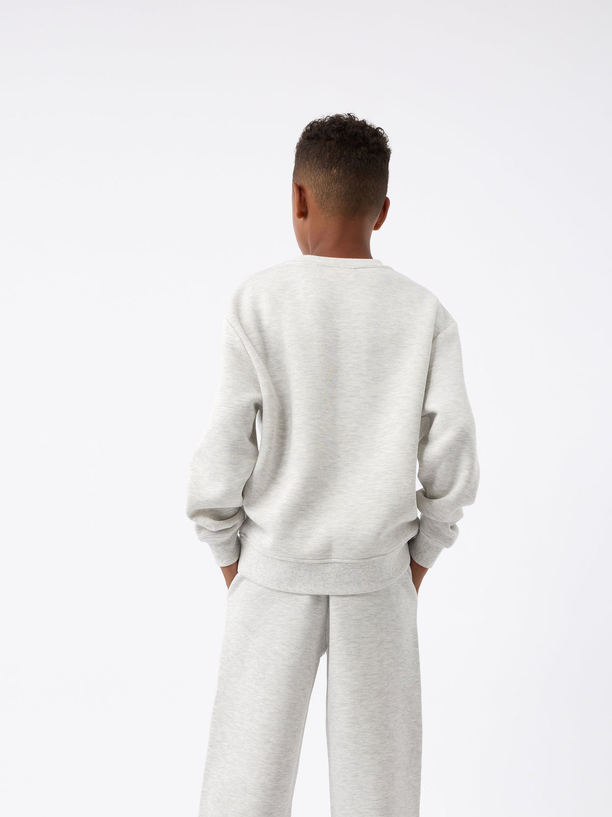 A child dressed in a light gray Kid's CityScape Crewneck from Cozy Earth, accompanied by matching pants, stands with their back to the camera. Their hands are tucked into their pockets against a plain white background. |Color:Heather Grey