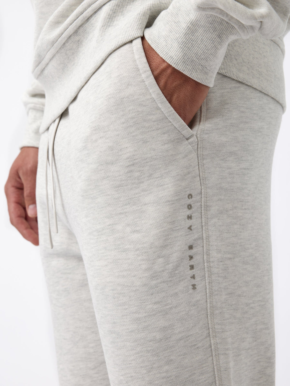 A person wearing Cozy Earth's Men's CityScape Jogger in light gray with their hand in the pocket. The joggers feature a subtle logo or text running vertically down the side and the fabric appears soft and comfortable. |Color:Heather Grey