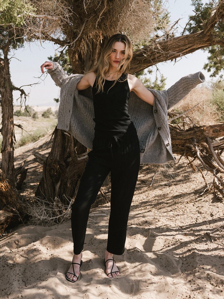 A woman stands on sandy ground under a large tree, wearing Cozy Earth’s Women’s Sunset Cropped Pant, a black tank top, sandals, and a gray cardigan draped over her shoulders as sunlight filters through the branches. |Color:Jet Black