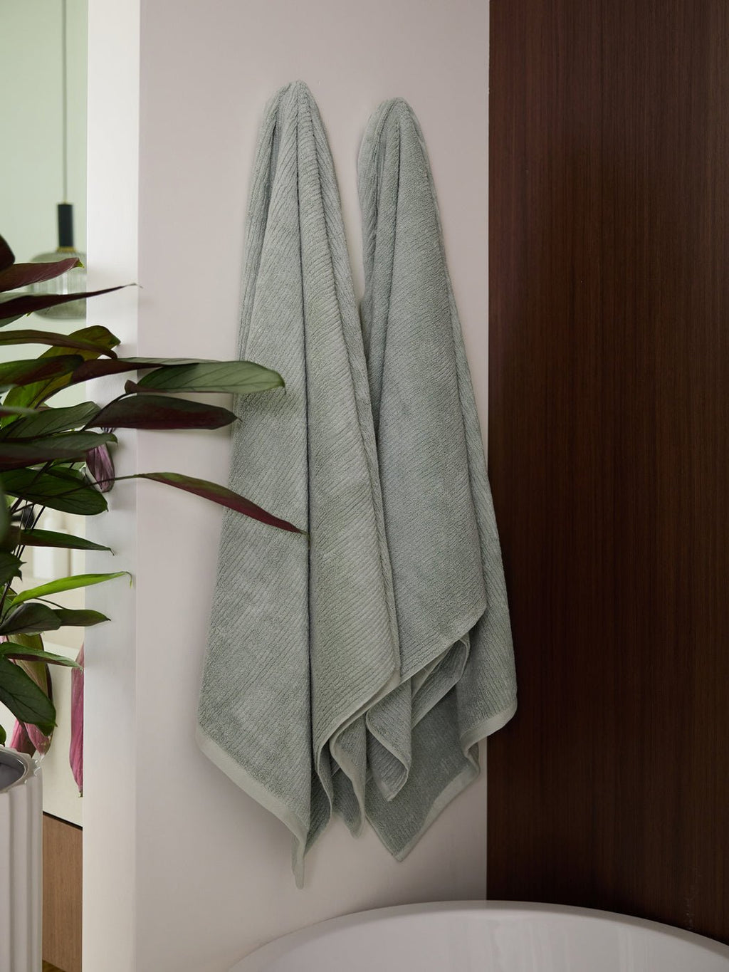 Two Cozy Earth Ribbed Terry Bath Towels in light gray hang side by side on a white wall near a wooden panel, with a green potted plant and part of a bathtub visible in the modern bathroom.|Color:Laurel
