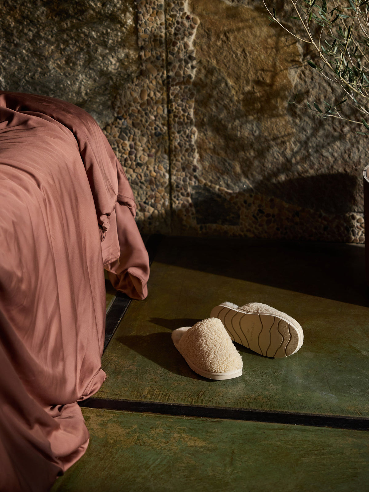 A pair of Cozy Earth Puffy Sheep Slippers are placed on a glossy, green floor next to a bed covered with a rust-colored, silky blanket. The background features a textured stone wall and part of a potted plant. |Color:Natural