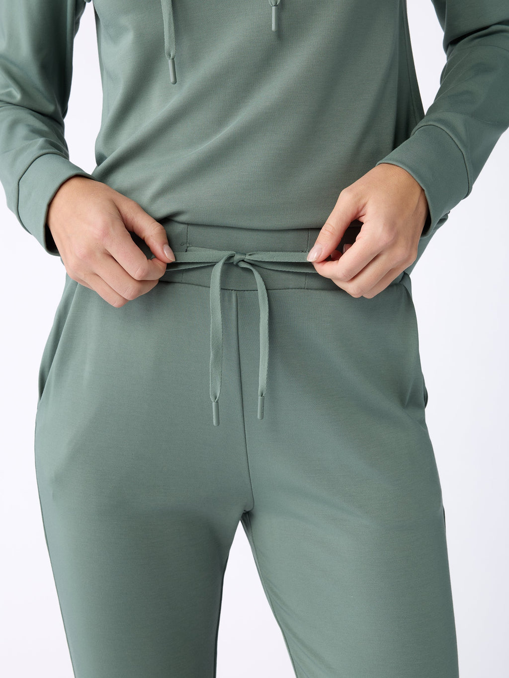 A person wearing the Women's Bamboo Jogger Pant TALL in green from Cozy Earth adjusts the drawstring on the pants. The image is focused on the torso and hands, highlighting the comfortable, casual outfit. |Color:Oakmoss