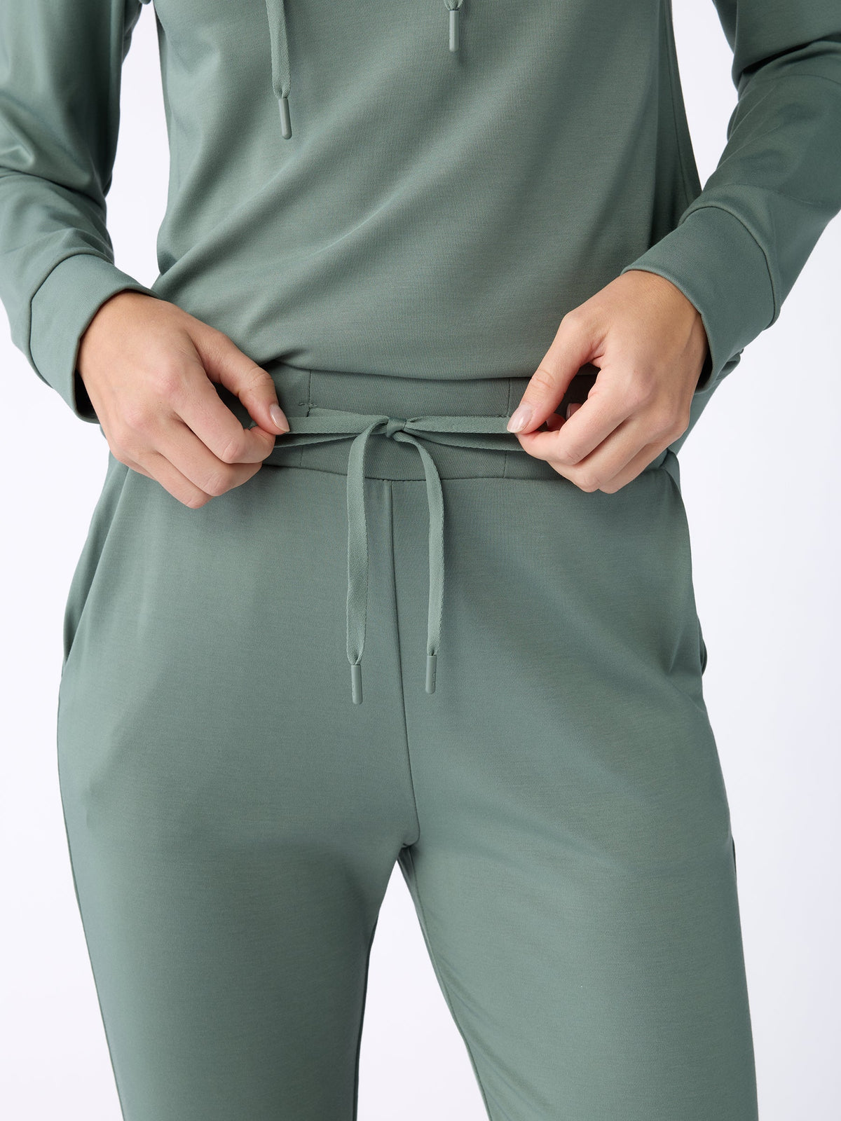 A person wearing the Women's Bamboo Jogger Pant TALL in green from Cozy Earth adjusts the drawstring on the pants. The image is focused on the torso and hands, highlighting the comfortable, casual outfit. |Color:Oakmoss
