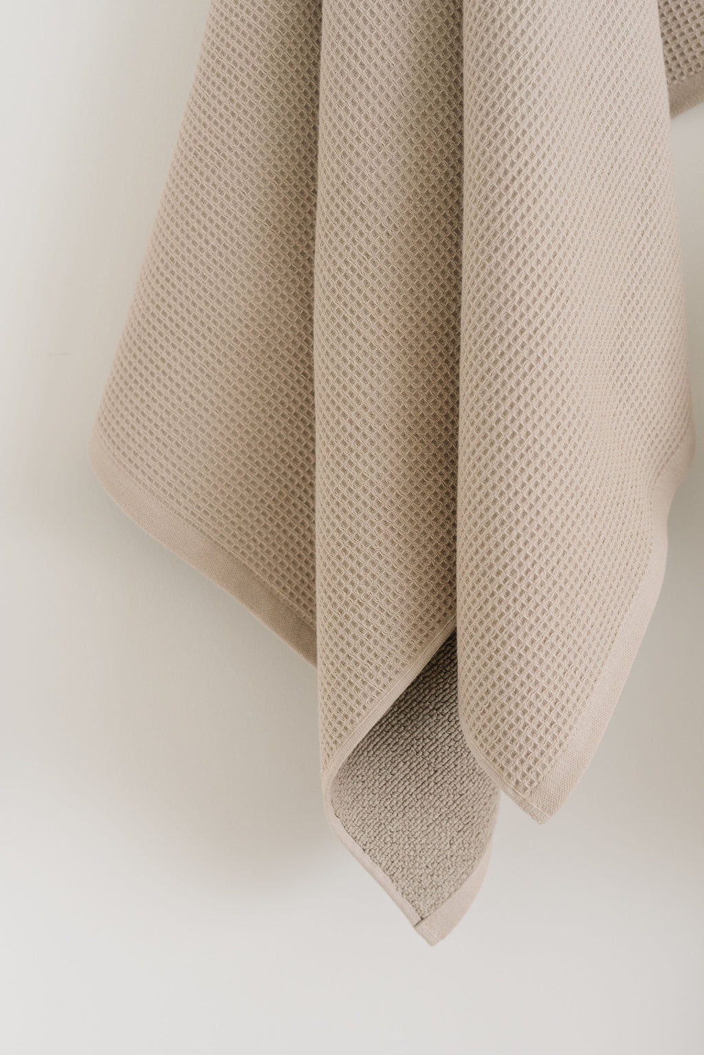 Waffle Bath Towel in the color Sand. Photo of Sand Waffle Bath Towel taken hanging from a towel hook in a white bathroom. The upper half of the towel and towel hook is not seen. |Color:Sand