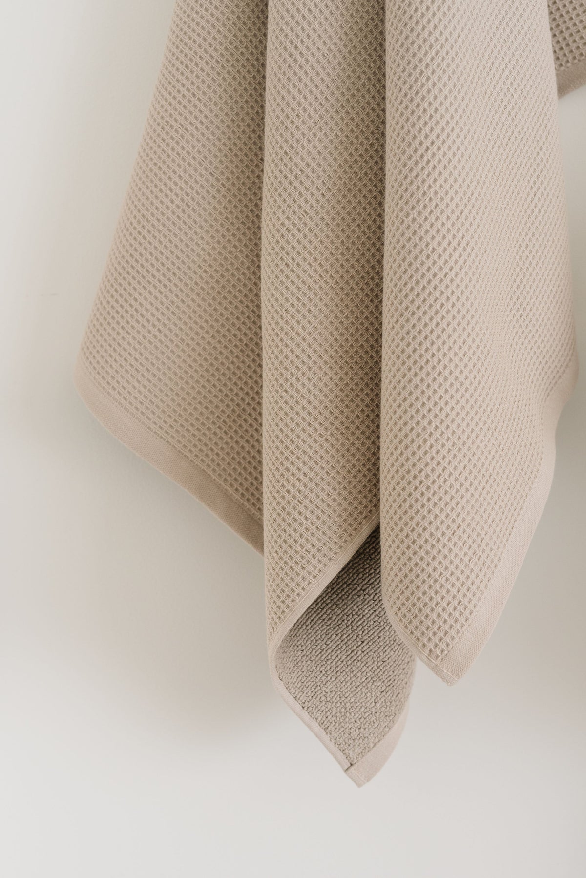 Waffle Bath Towel in the color Sand. Photo of Sand Waffle Bath Towel taken hanging from a towel hook in a white bathroom. The upper half of the towel and towel hook is not seen. |Color:Sand