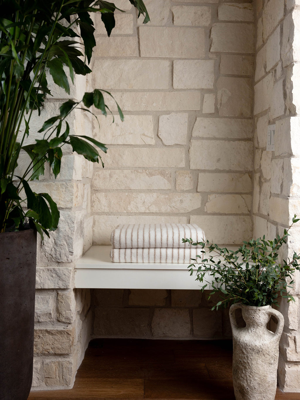 A cozy nook with a white stone wall features a minimalist cream-colored bench topped with two Luxe Bath Towels from Cozy Earth. The space is adorned with two lush green plants, one in a rustic gray pot and the other in an earthenware vase. Natural light highlights the serene setting. |Color:Sand Stripe