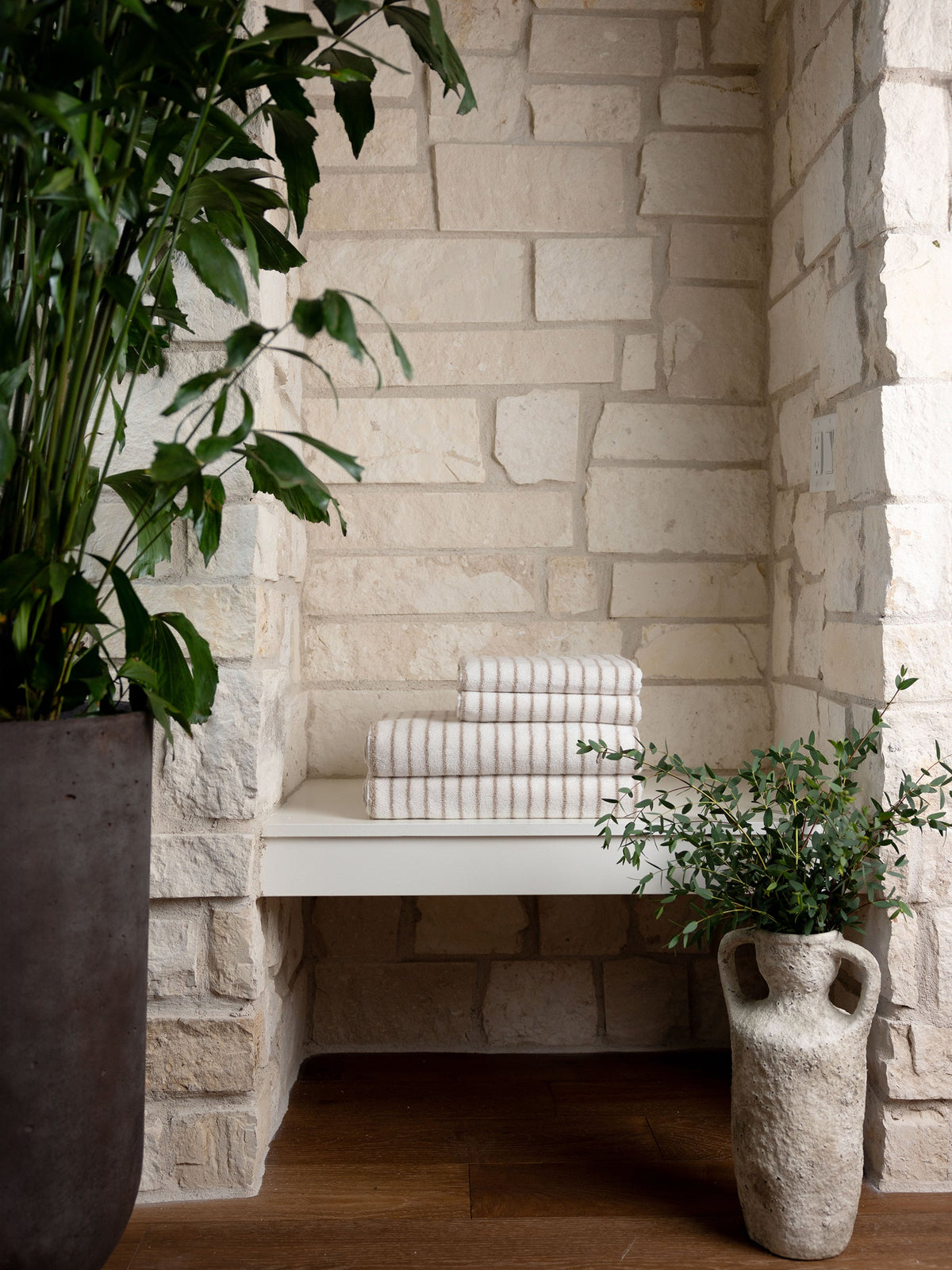 A serene corner with Luxe Hand Towels from Cozy Earth neatly stacked on a white floating shelf, surrounded by large potted plants. Walls feature light natural stone, and the floor is of dark wood, creating a harmonious blend of textures and natural colors. |Color:Sand Stripe