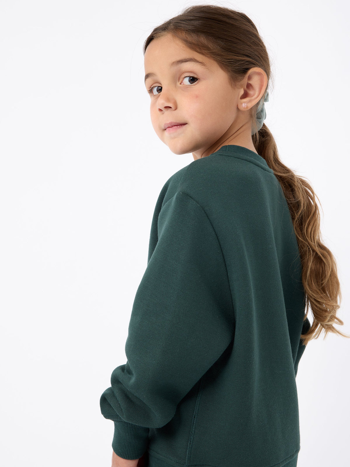 A young girl with long brown hair in a ponytail, wearing the Cozy Earth's Kid's CityScape Crewneck, looks over her shoulder against a plain white background. |Color:Spruce