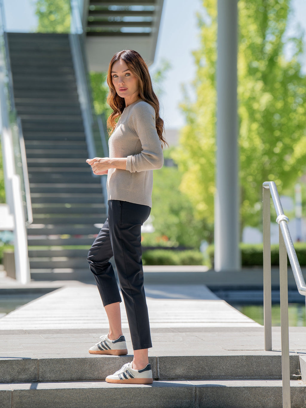 A woman with long brown hair stands on an outdoor staircase, wearing a beige Women's Classic Crewneck by Cozy Earth, black pants, and gray Adidas sneakers. She looks over her shoulder with a serious expression. The background features a modern structure and green trees. |Color:Taupe