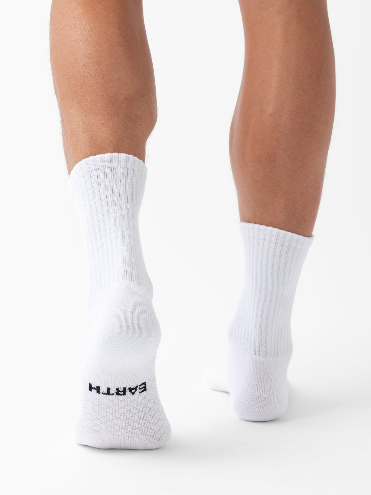 A person stands on tiptoe, showing the back of their legs and feet in white ribbed Essential Calf Socks by Cozy Earth. "EARTH" is printed in black on the bottom of the left sock. The background is white. |Color:White