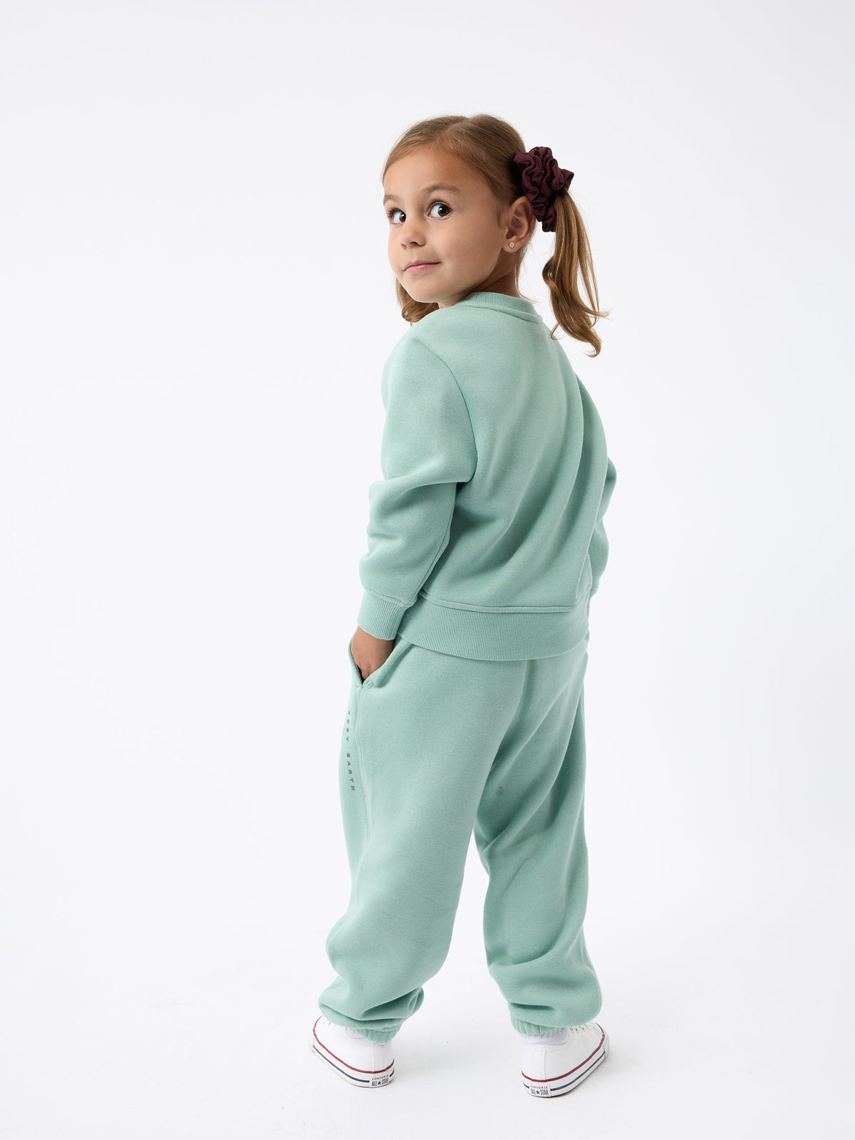 A young girl stands facing away, glancing back with a smile. She is dressed in a Kid's CityScape Crewneck by Cozy Earth paired with white sneakers, and her hair is styled in two pigtails secured with hair ties. The backdrop is white and minimalistic. |Color:Wintergreen