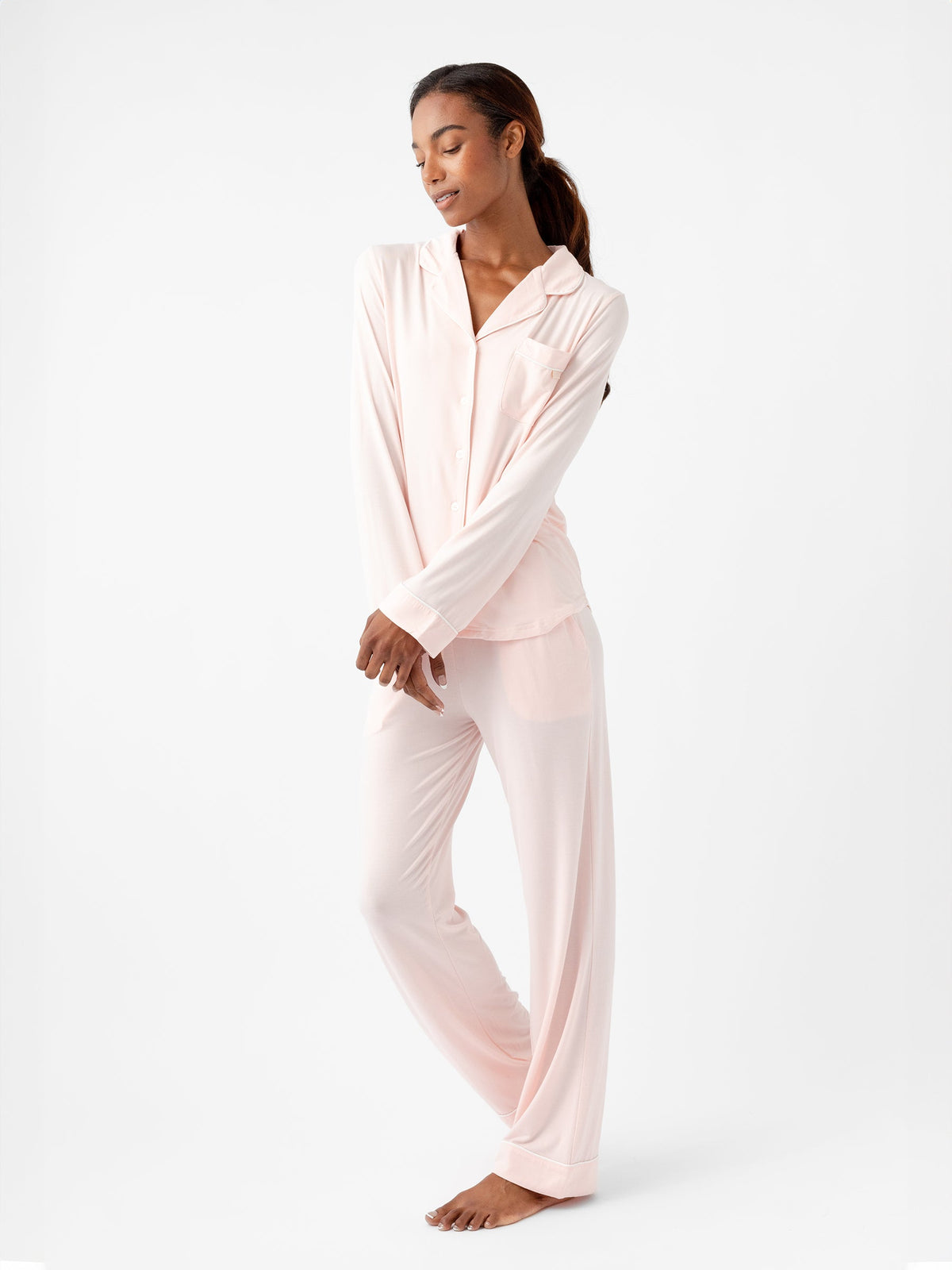 A woman stands barefoot against a plain white background, wearing the Women's Long Sleeve Bamboo Pajama Top in Stretch-Knit TALL from HIDE, in light pink. The pajama top has a collar and buttons, and the matching pants are loose-fitting. She has her arms folded slightly and looks down with a relaxed expression. |Color:Blossom