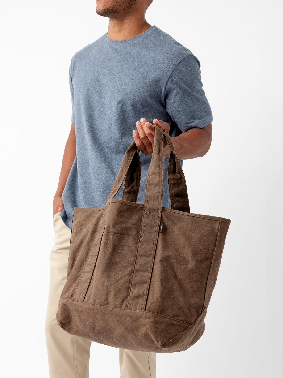 A person in a blue t-shirt and beige pants holds a large Waxed Canvas Tote by Cozy Earth. The tote bag has long straps and appears spacious. The background is plain white. The person's face is not visible. |Color:Cedar