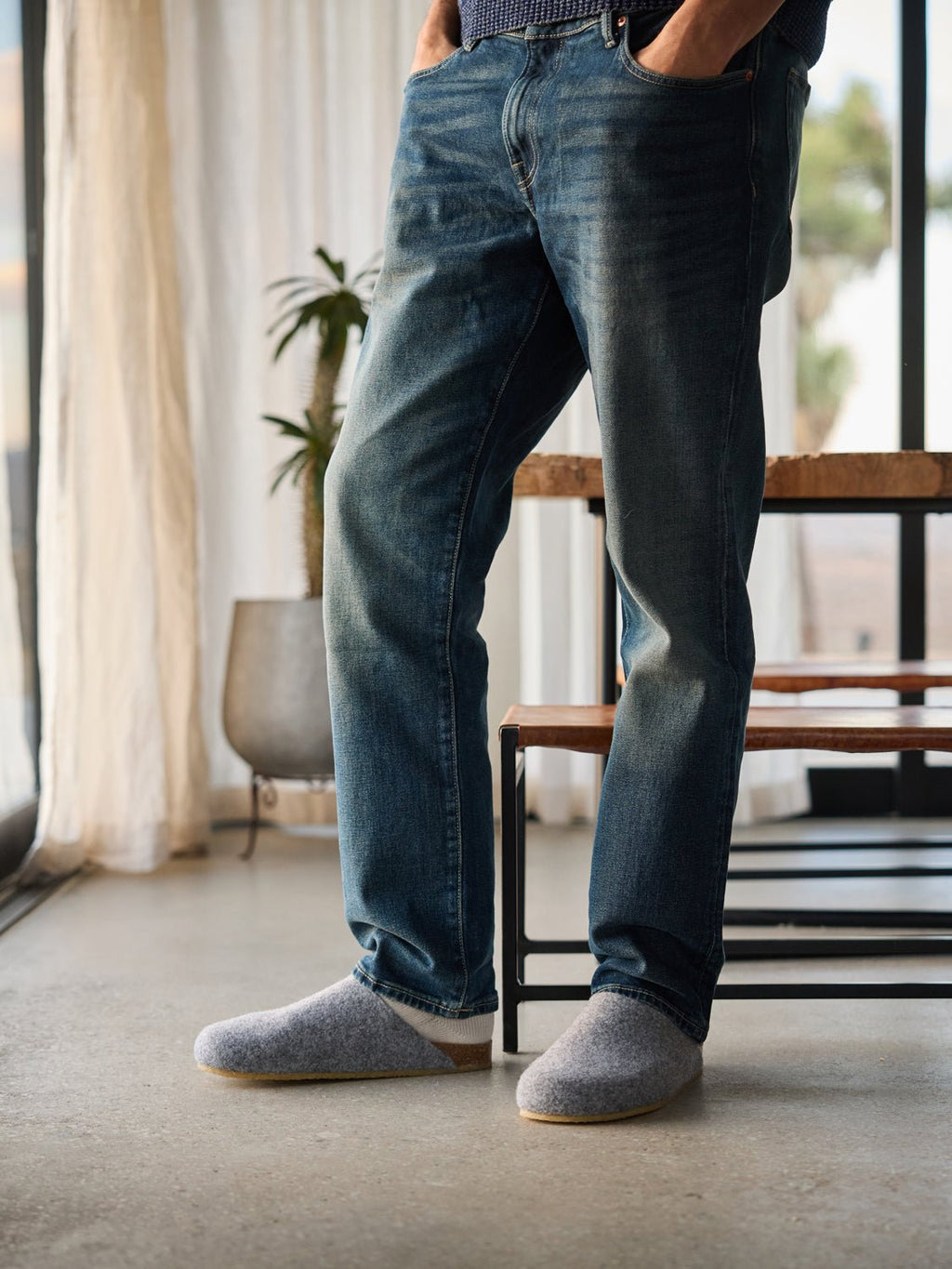A person in blue jeans and Cozy Earth's gray Lakehouse Clogs stands in a sunlit room with hands in pockets. A wooden bench, a potted plant, and sheer curtains are visible in the background. |Color:Charcoal
