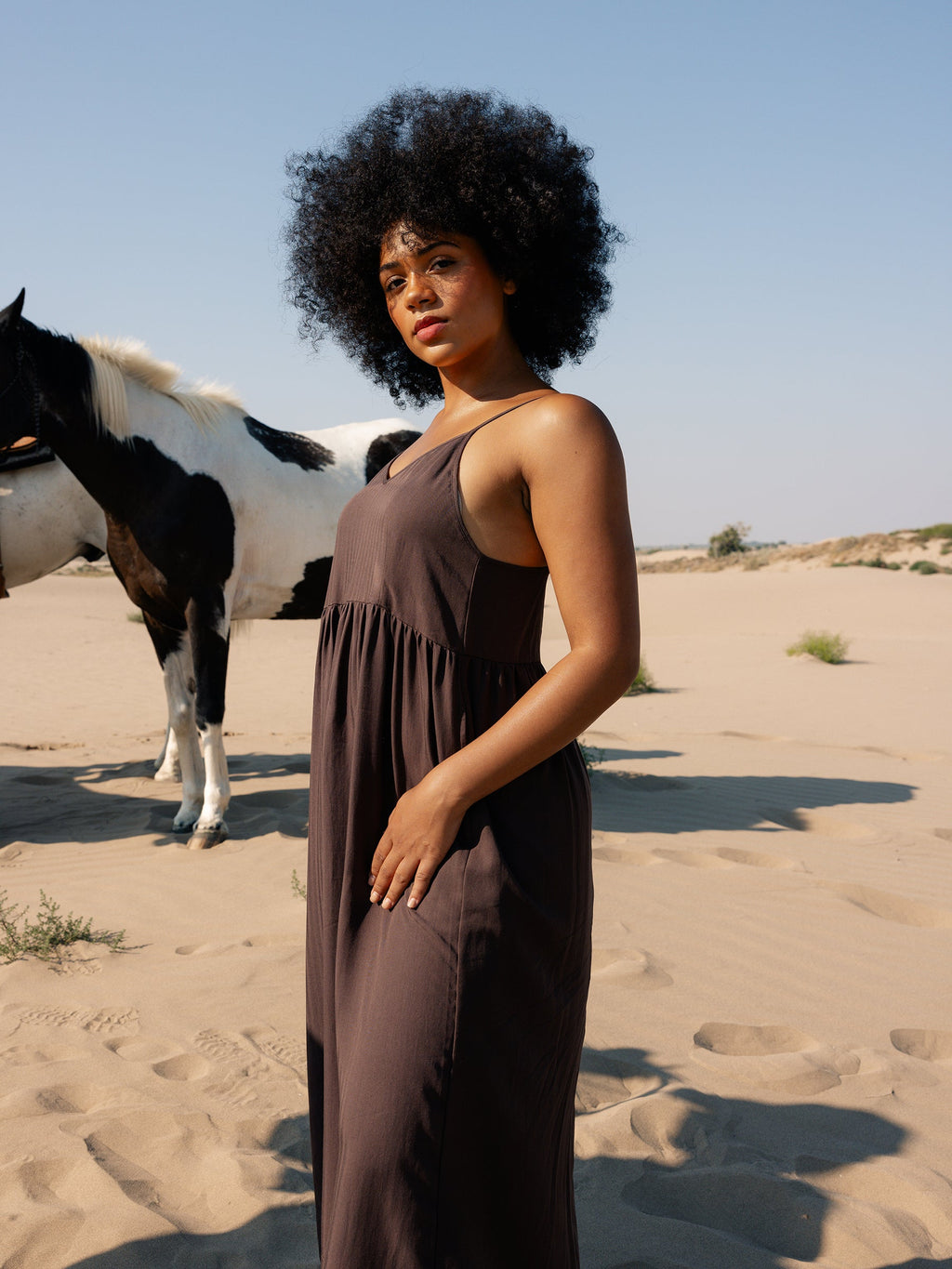 A woman with curly hair wears a loose, dark Cozy Earth Women's Sunset Gathered Dress, standing in a sandy desert landscape. A black and white horse stands in the background under a clear blue sky. |Color:Cacao