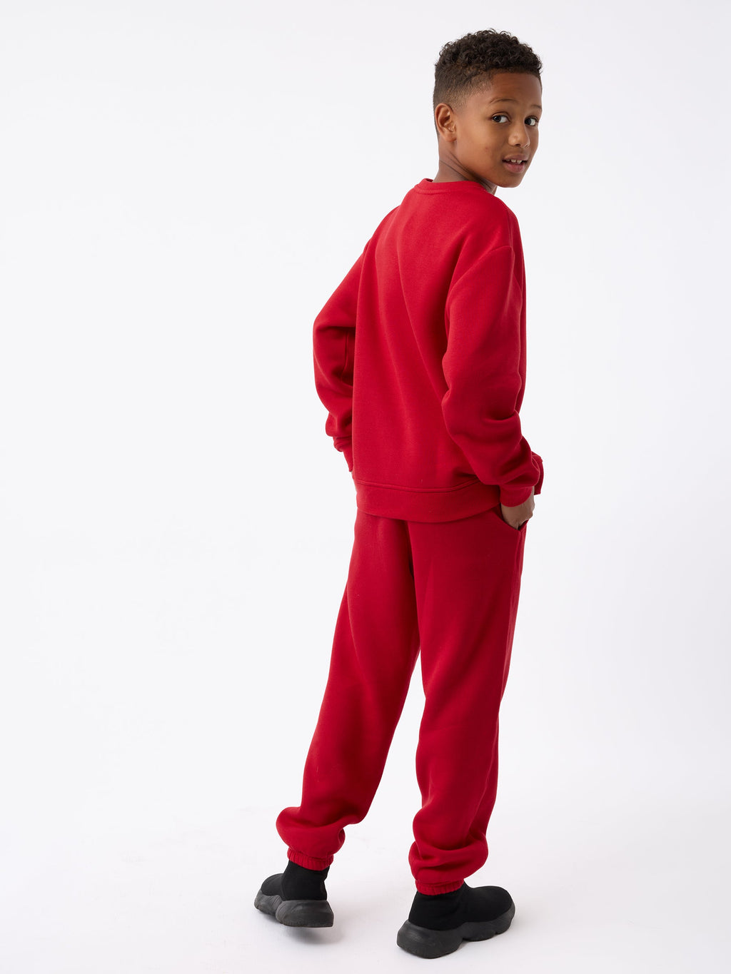 A young boy wearing the Kid's CityScape Crewneck by Cozy Earth and black shoes stands with his back turned, looking over his shoulder and smiling against a plain white background. |Color:Crimson