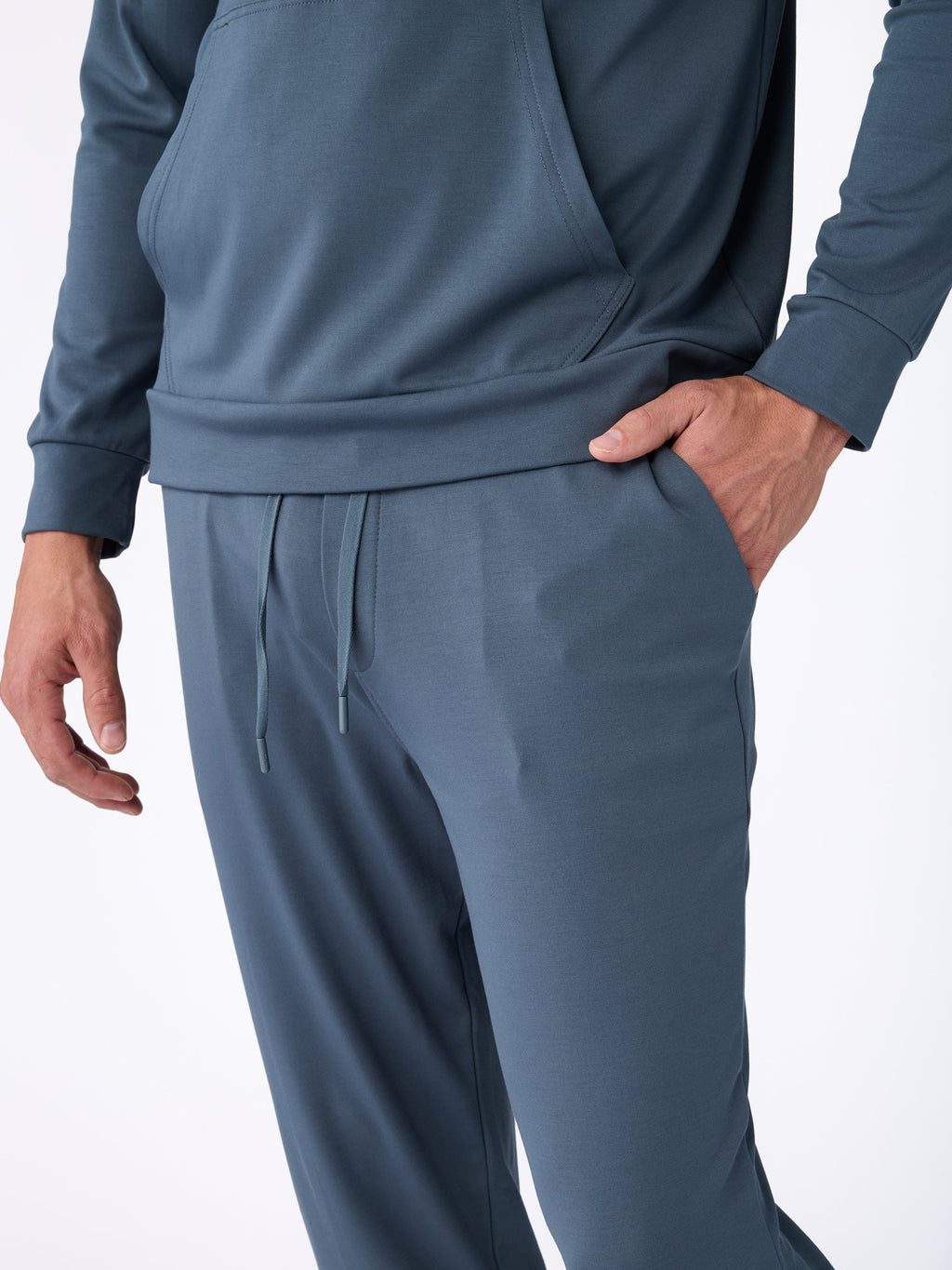 A person is dressed in the Men's Ultra-Soft Bamboo Pullover Crew by Cozy Earth, featuring a blue-gray color. Their right hand is tucked into the pocket of the pants, while the top sports long sleeves and a front pouch pocket against a plain white background. |Color:Dawn