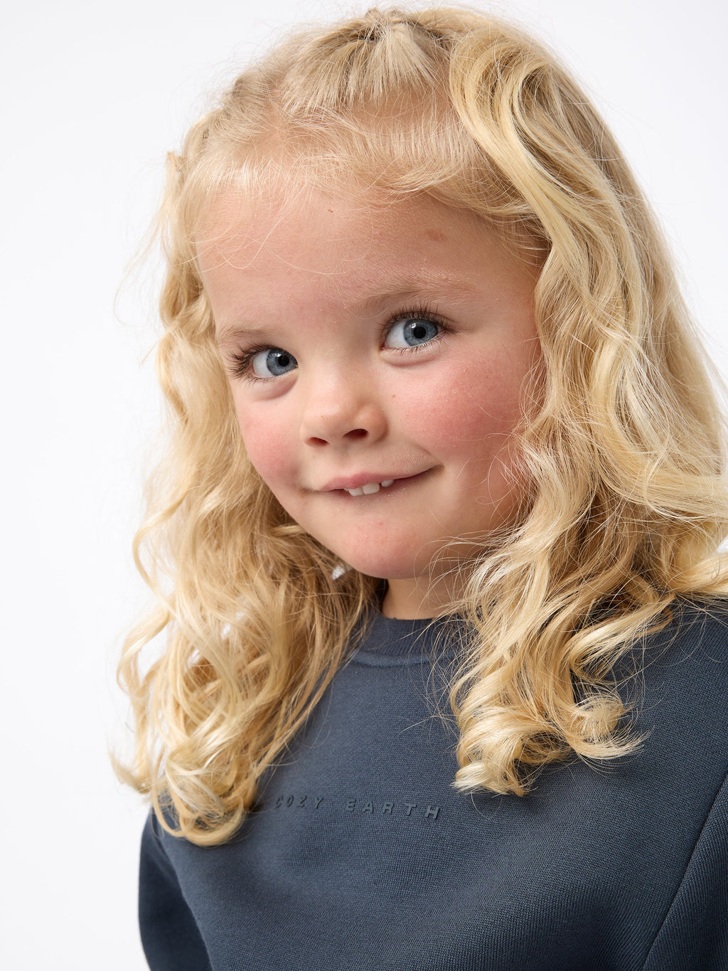 A young child with curly blonde hair smiles slightly, wearing the Kid's CityScape Crewneck from Cozy Earth, featuring a dark gray design with subtle text. The background is plain white. |Color:Eclipse