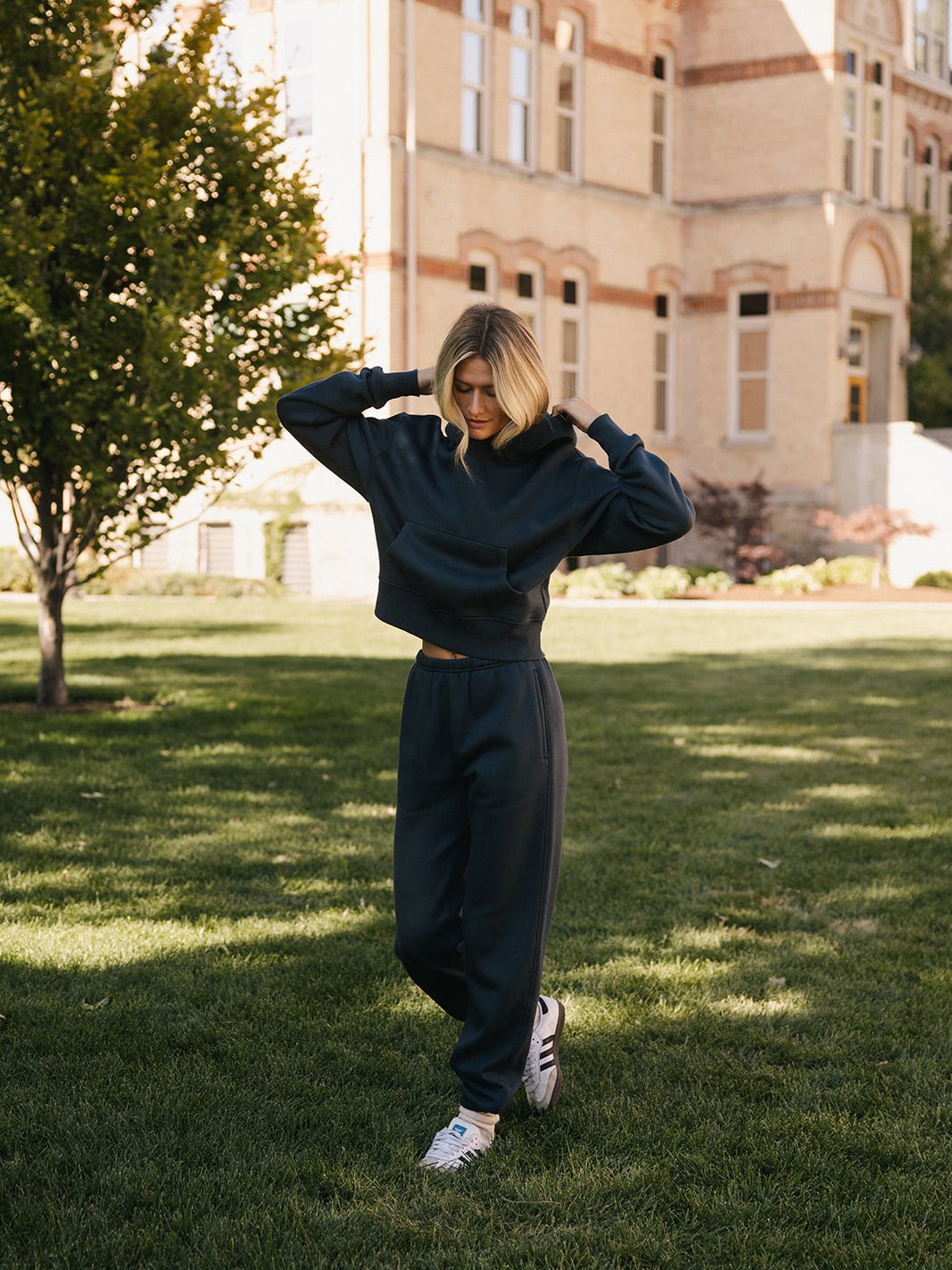 A person with shoulder-length blonde hair is standing on grass in front of a large brick building with tall windows. They are wearing a dark CityScape Cropped Hoodie by Cozy Earth, matching pants, and white sneakers, with their hands raised to adjust their hair or clothing. |Color:Eclipse